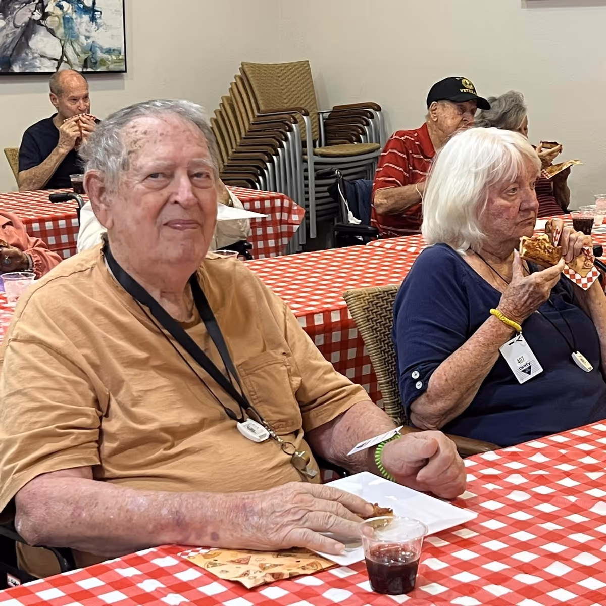 Several elderly people sitting at tables covered with red and white checkered tablecloths, eating pizza and drinking beverages in a communal dining area.
