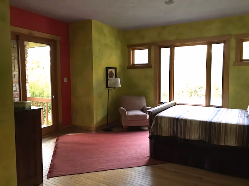 Bright bedroom with striped bedding, armchair, lamp, large windows, and a red area rug