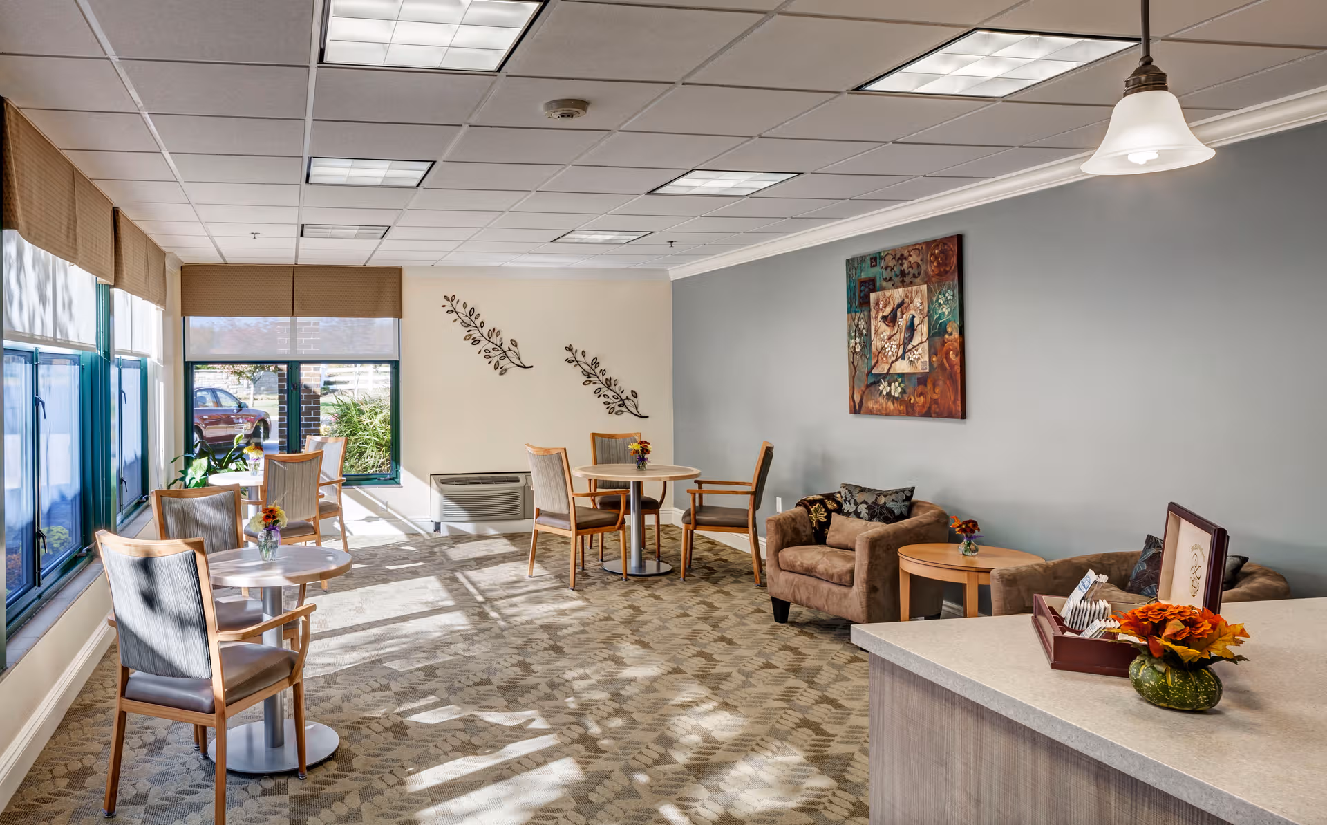Bright, airy lounge with tables, chairs, a couch, and wall art in a senior living center.