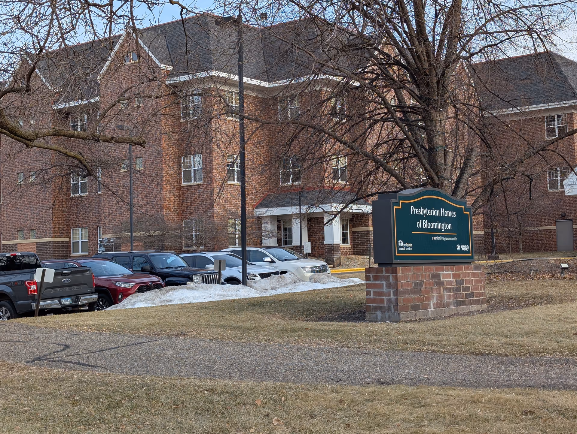 Brick senior living building with parked cars and a prominent sign reading "Presbyterian Homes of Bloomington" on the lawn.