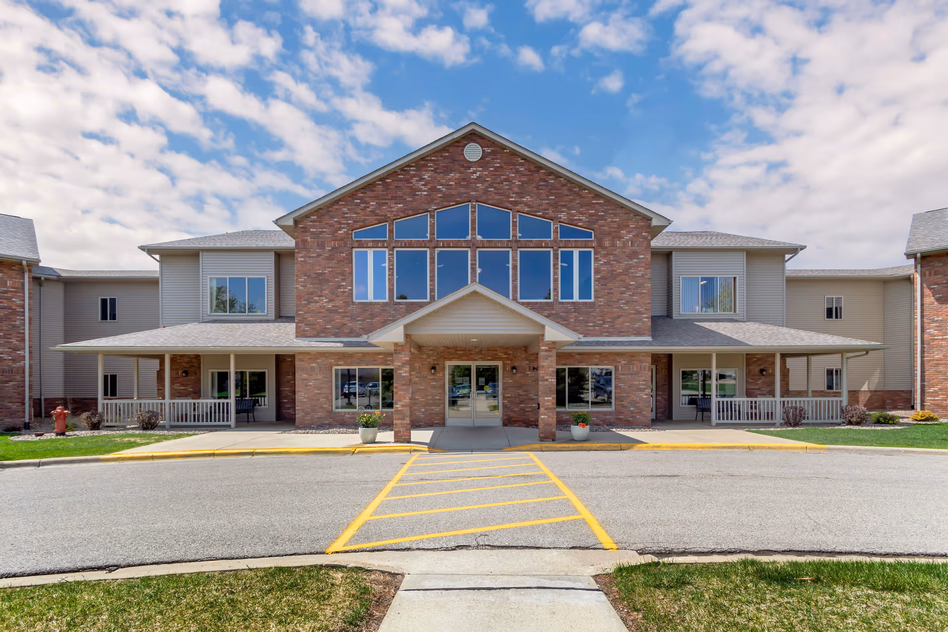 Front exterior view of a two-story senior living facility building with a brick facade and large windows under a partly cloudy sky. There is a covered entrance with two pillars, a driveway with yellow painted lines, and some greenery on either side.