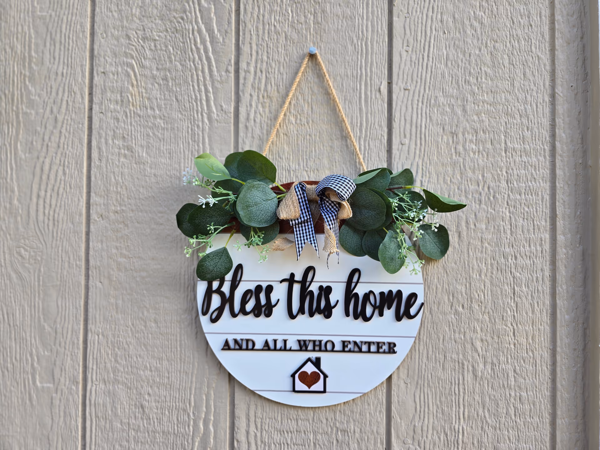 Decorative hanging sign reading 'Bless this home' with greenery on a wooden wall.