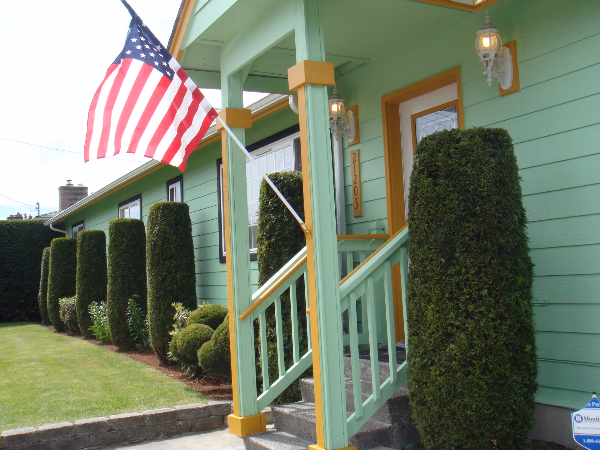 Mint-green building with front porch, American flag, and tall manicured hedges.
