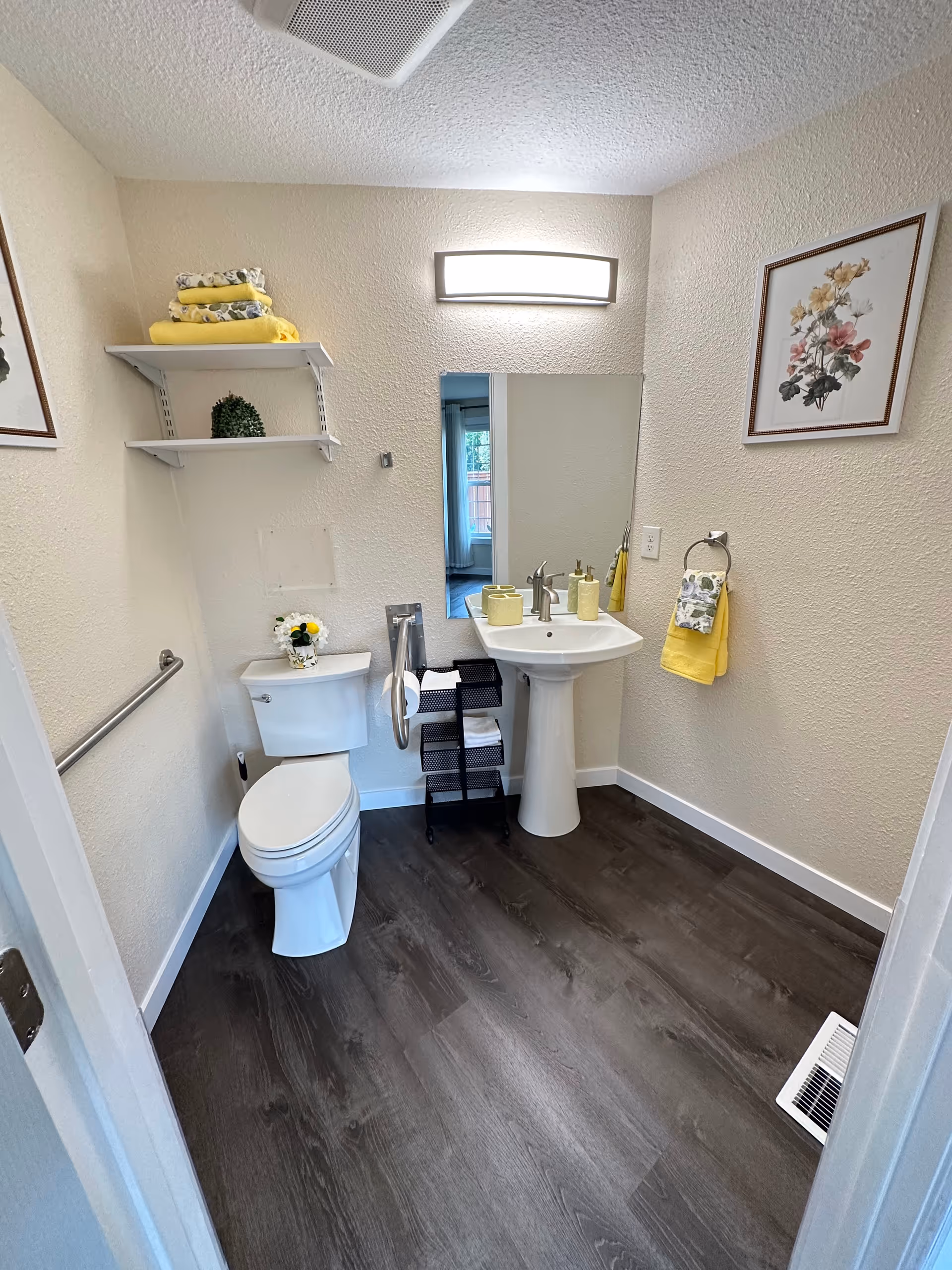 Small beige bathroom with toilet, pedestal sink, mirror, shelves, and yellow decor.