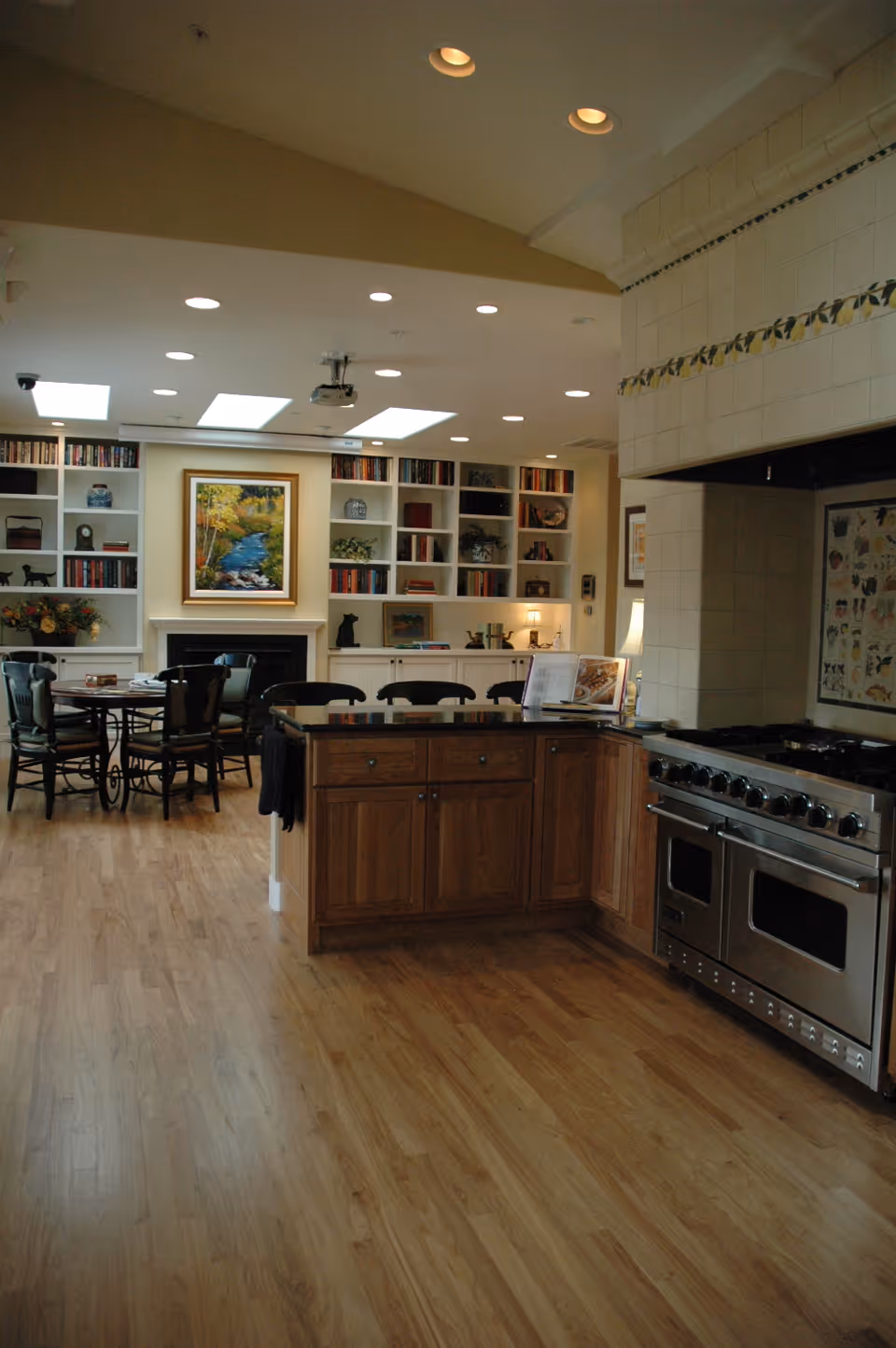 A spacious kitchen with wooden cabinets and a large stainless steel stove on the right. The kitchen opens into a dining area with a round table and chairs. In the background, there is a white built-in bookshelf filled with books and decorative items, and a framed painting of a river scene above a fireplace. The ceiling has recessed lighting and skylights.