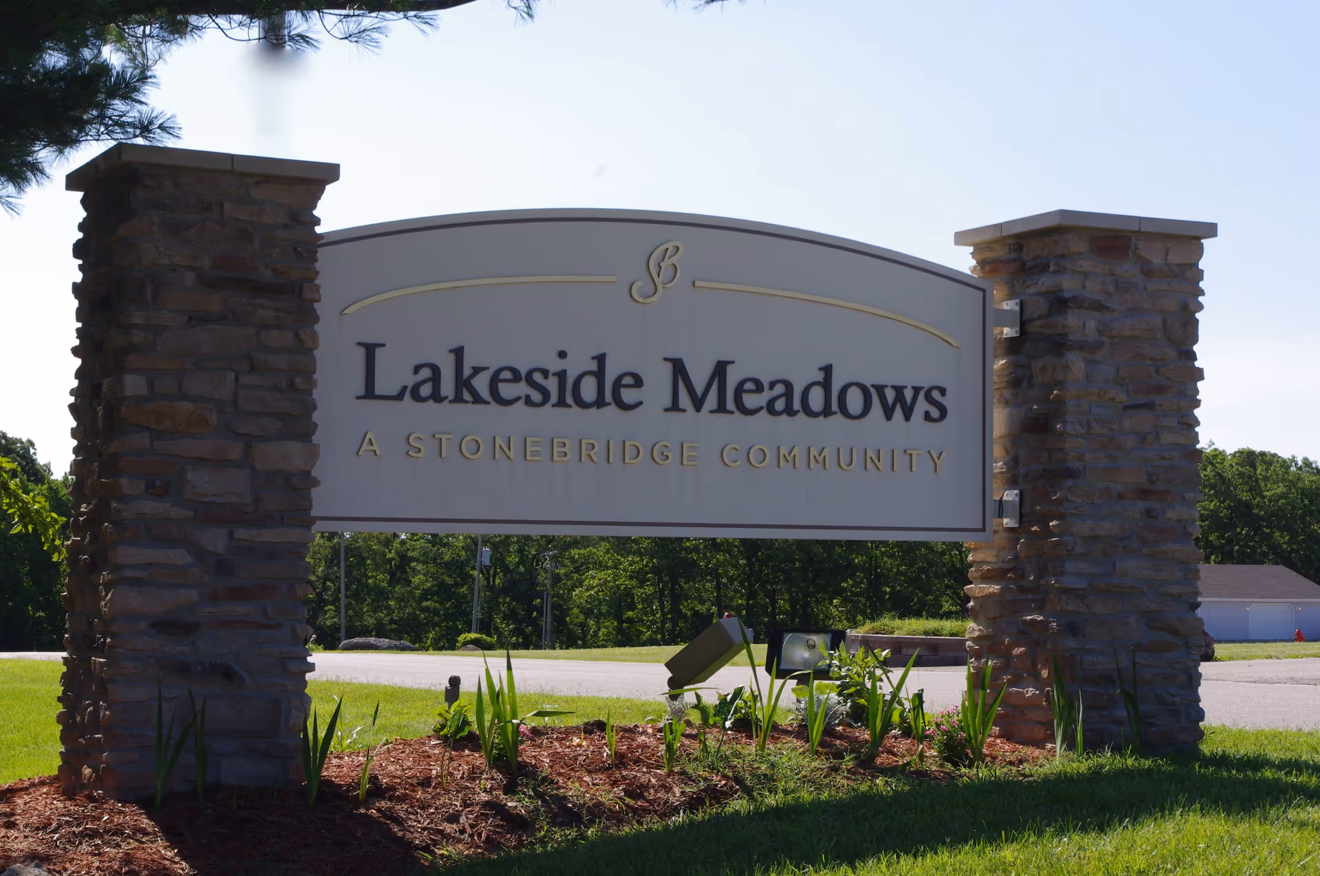 Stone sign for Lakeside Meadows, a Stonebridge community, mounted between two stone pillars with grass and plants in the foreground and trees in the background under a clear sky.