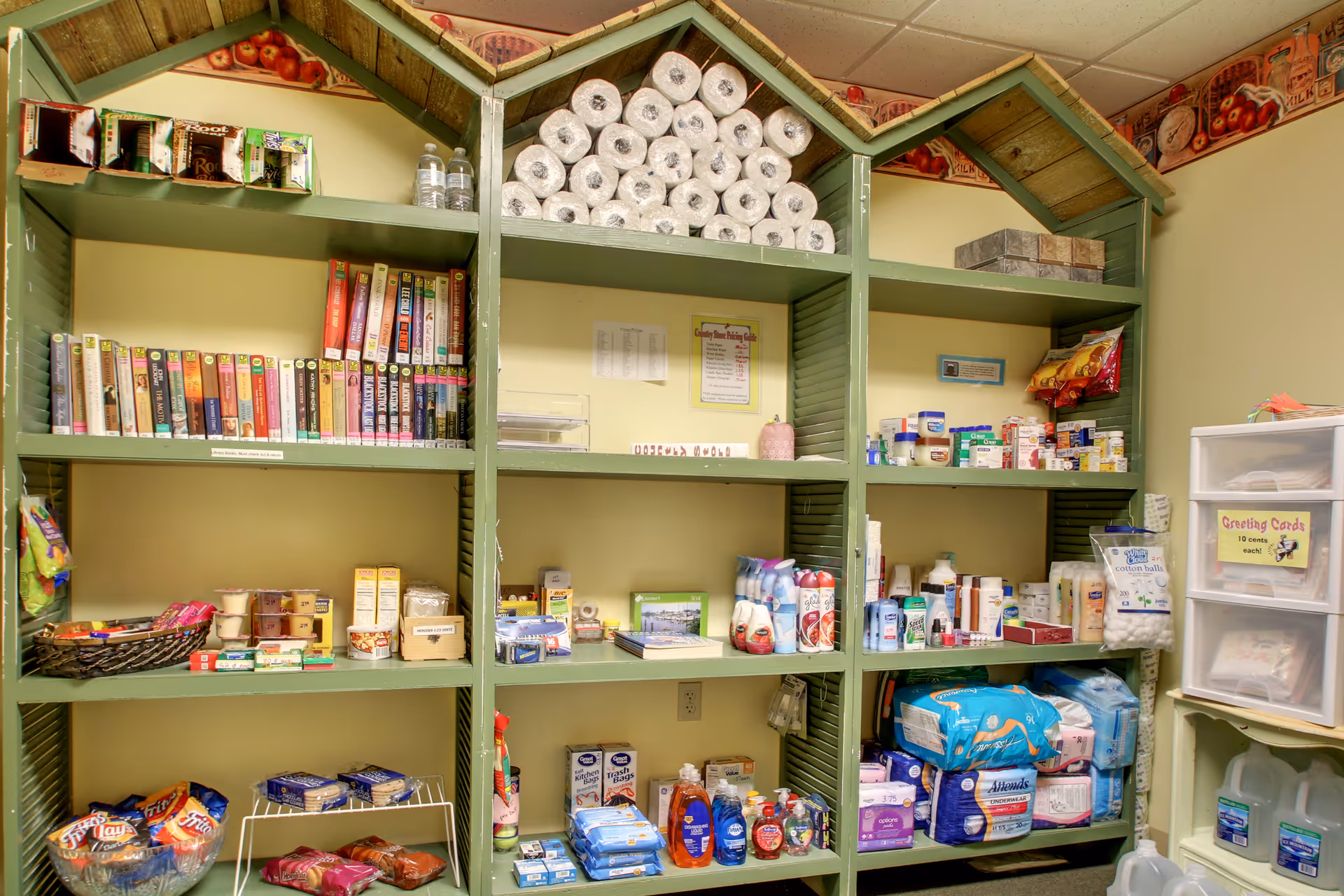 Shelves stocked with various items including books, paper towels, snacks, personal care products, and household supplies in a room with light yellow walls and a decorative border near the ceiling.