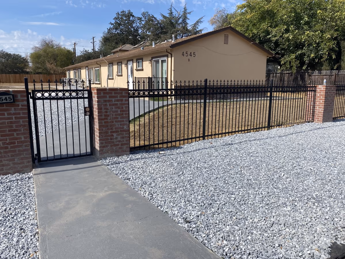 Single-story beige residential building marked "4545" behind a black metal fence and gated entrance with a gravel yard and concrete walkway.