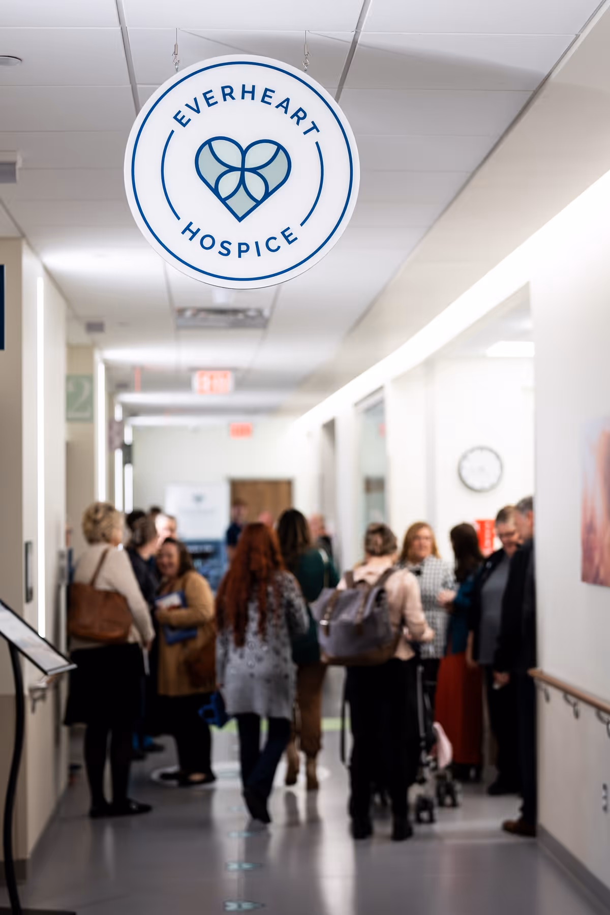 Round EverHeart Hospice sign hangs in a busy hallway with people and staff nearby.