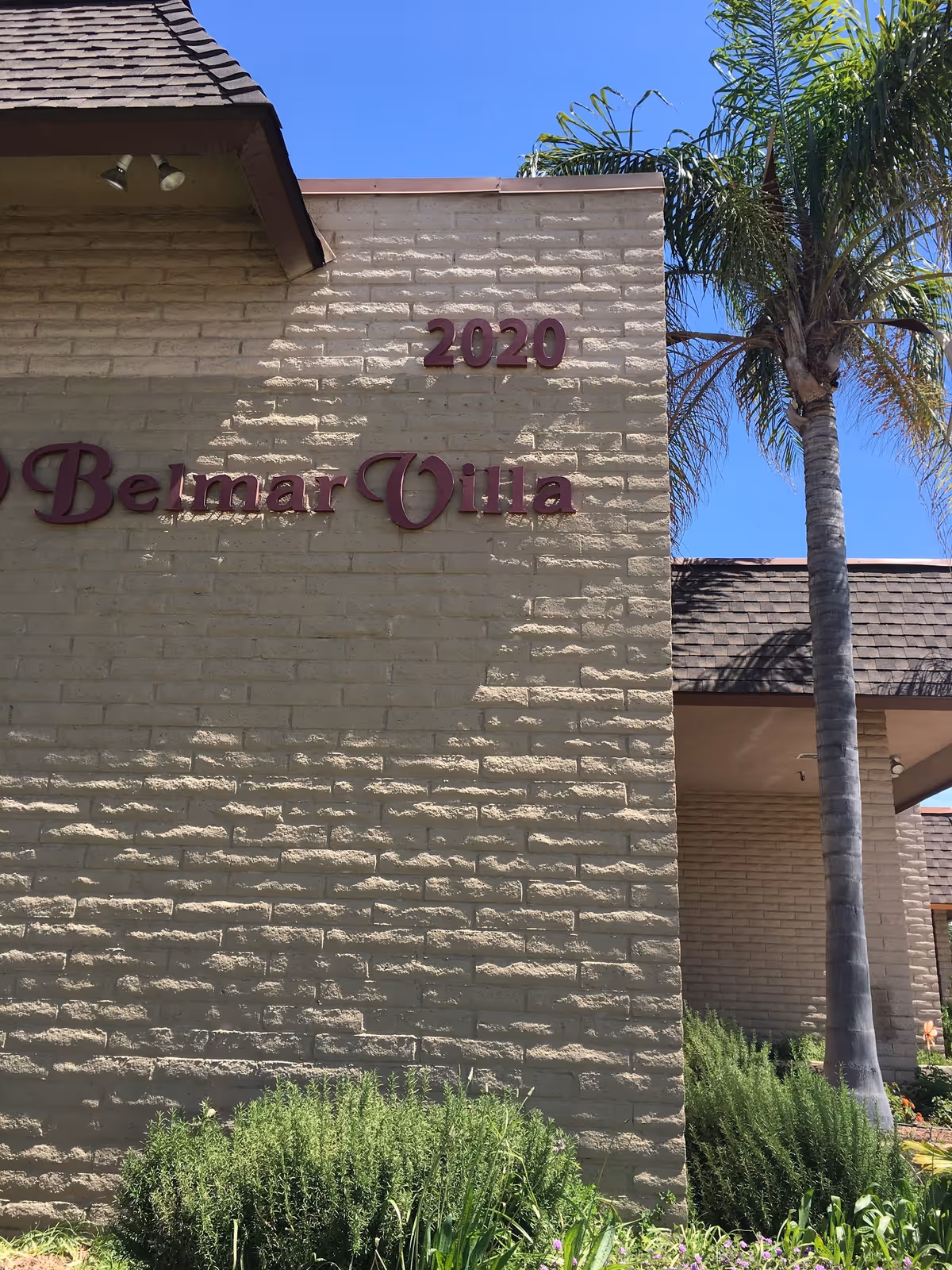 Front exterior of Belmar Villa with 2020 sign, beige brick wall, and a palm tree.
