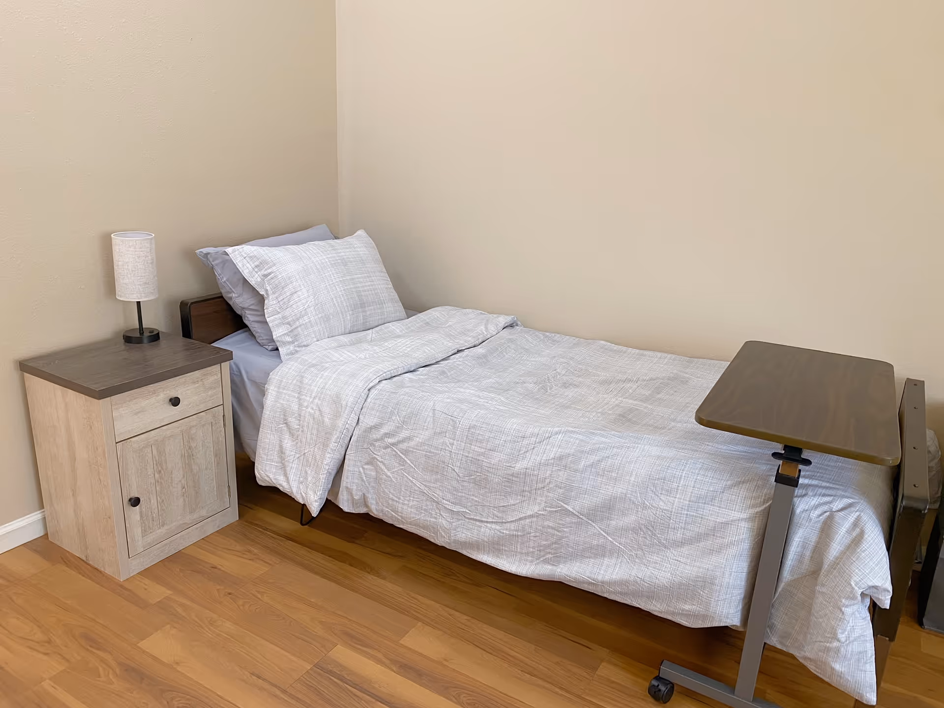 A compact bedroom with a bed, nightstand, lamp, and adjustable over-bed table.