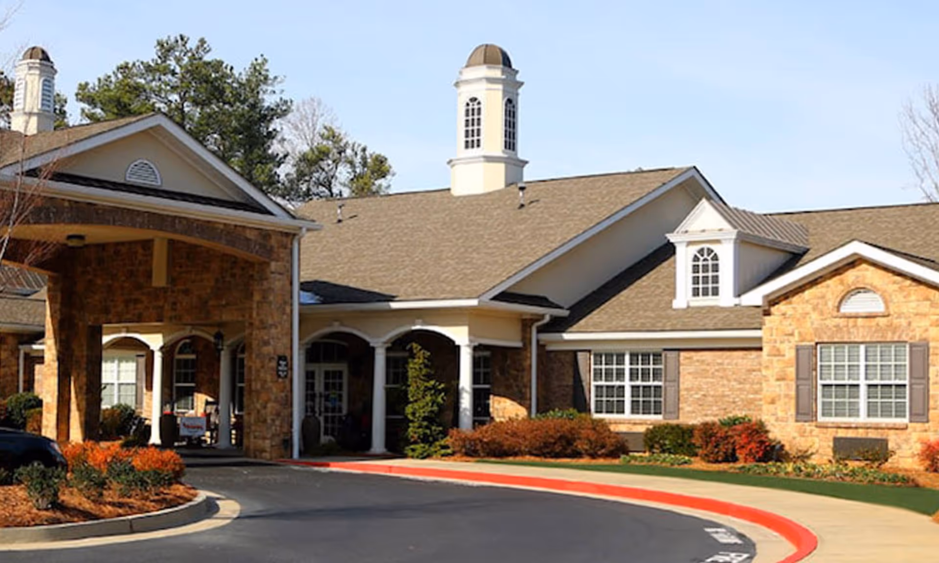 Exterior view of Addington Place of Johns Creek building with a covered entrance, stone and brick facade, multiple windows, and landscaped bushes and trees under a clear sky.
