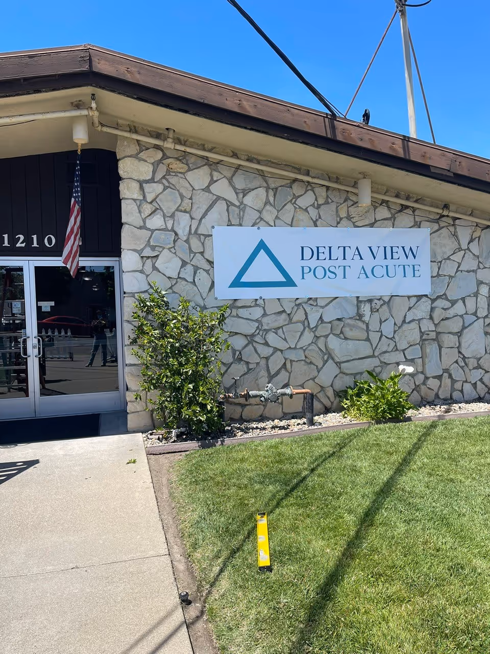 Stone facade building with Delta View Post Acute sign, entrance door, flag, and landscaping.