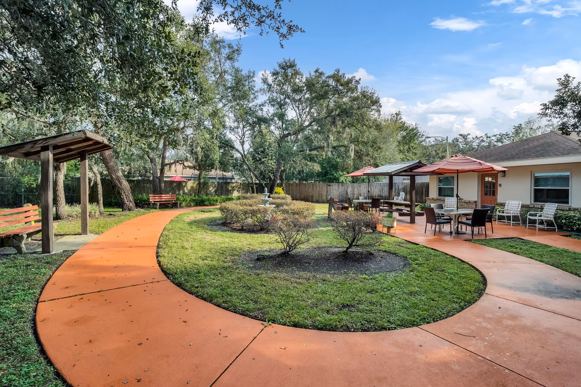 Outdoor garden area at Elevated Estates Forest Oaks featuring a curved orange walkway, green grass, bushes, trees, benches, and a seating area with tables, chairs, and red umbrellas near a building under a partly cloudy sky.