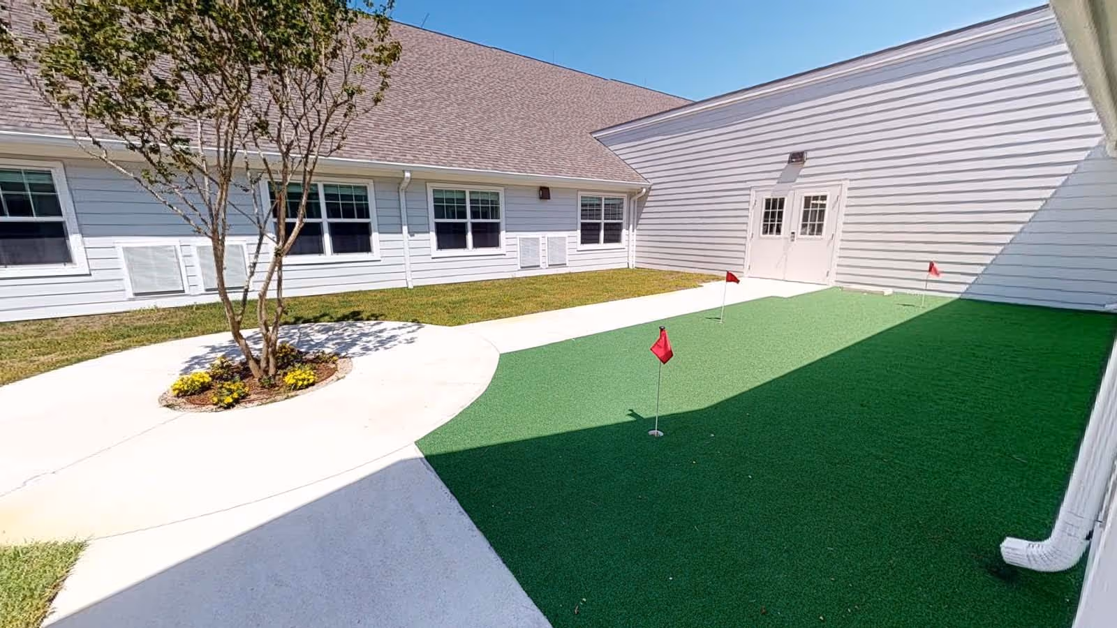 Outdoor courtyard area with a small artificial putting green featuring three red flags, surrounded by white buildings with multiple windows and a tree planted in a circular concrete planter.