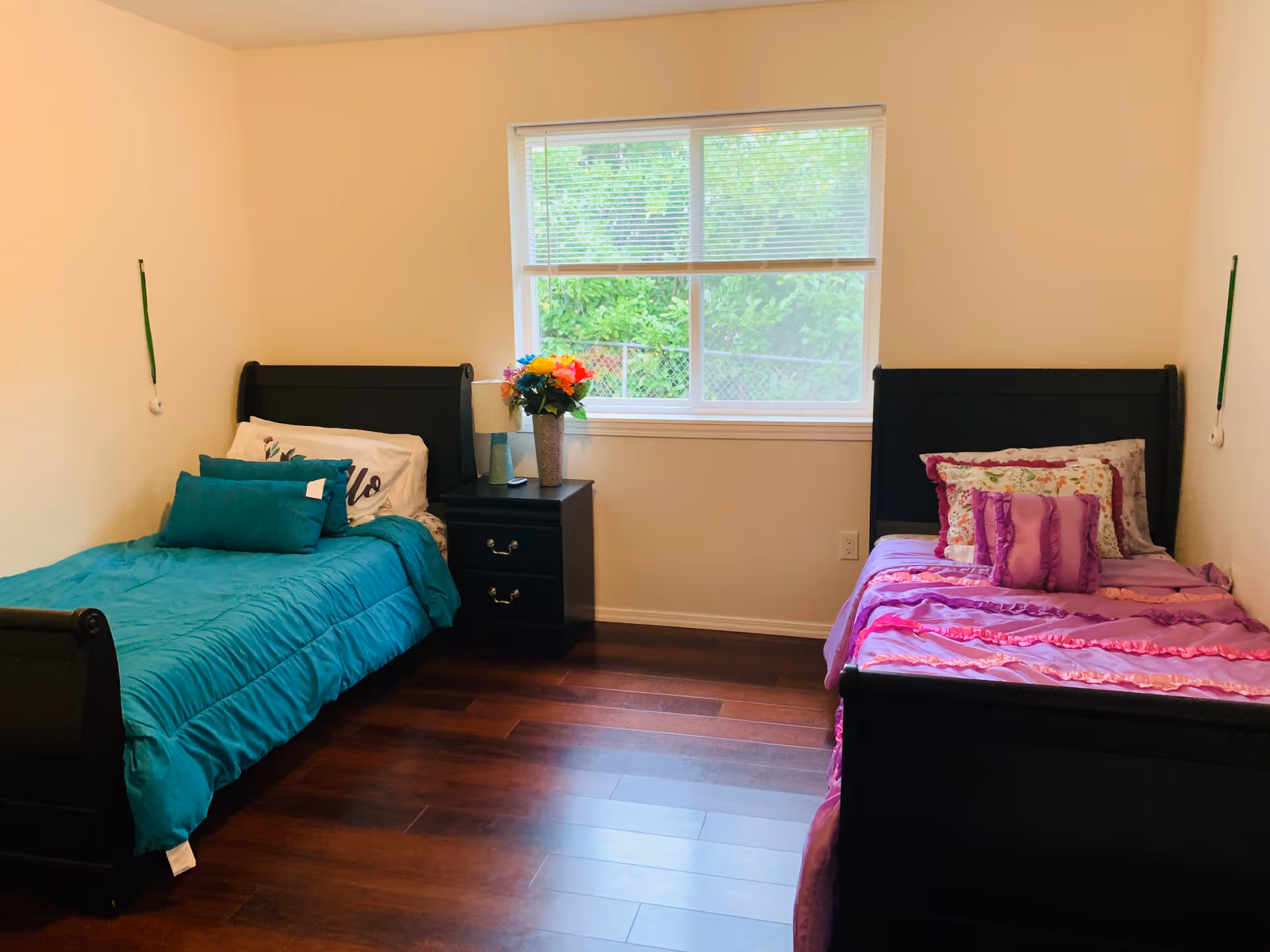 Two single beds with teal and pink bedding flank a nightstand with flowers by a window