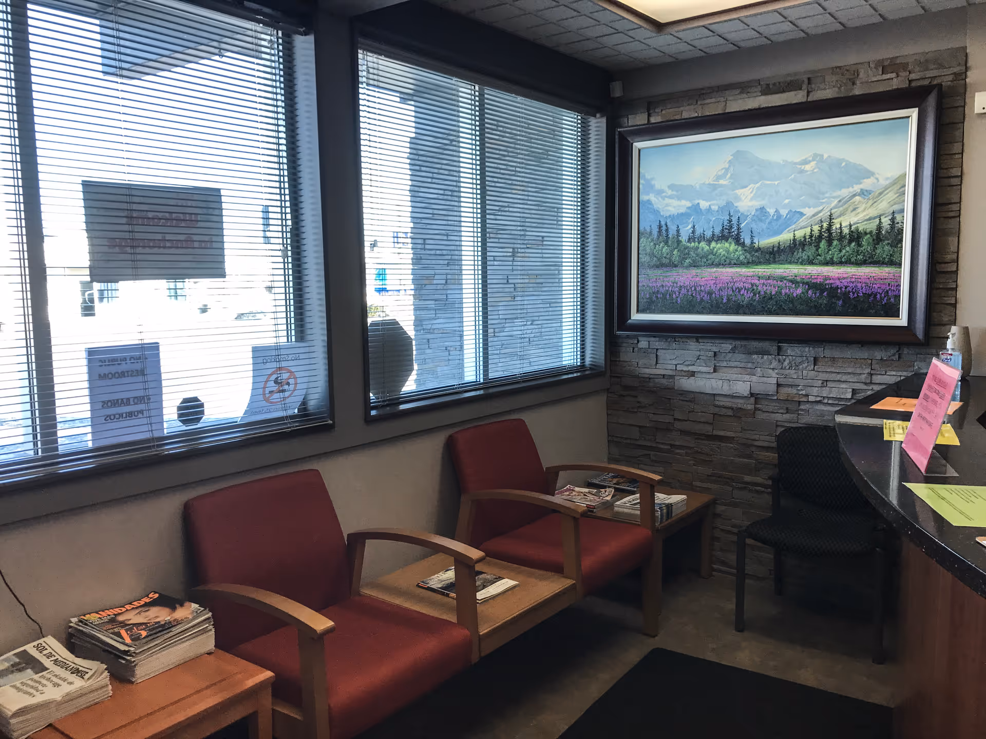 A waiting area with red chairs, magazines, large windows with blinds, and a framed landscape painting.
