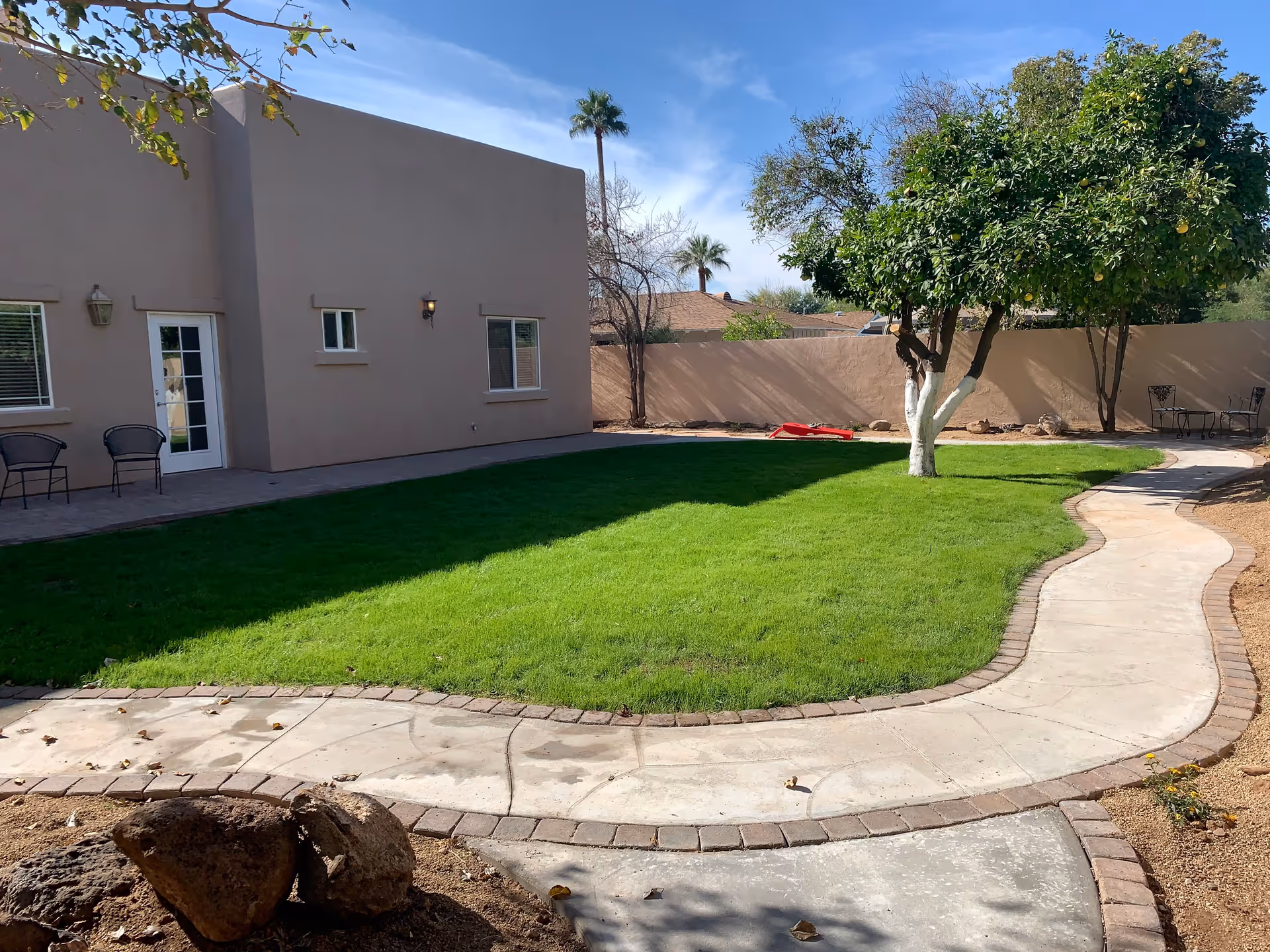 Outdoor garden area with a curved concrete pathway bordered by bricks, green grass lawn, a tree with a white-painted trunk, and a beige building with windows and a door. There are two black chairs near the building and a small table with two chairs in the background near the wall.