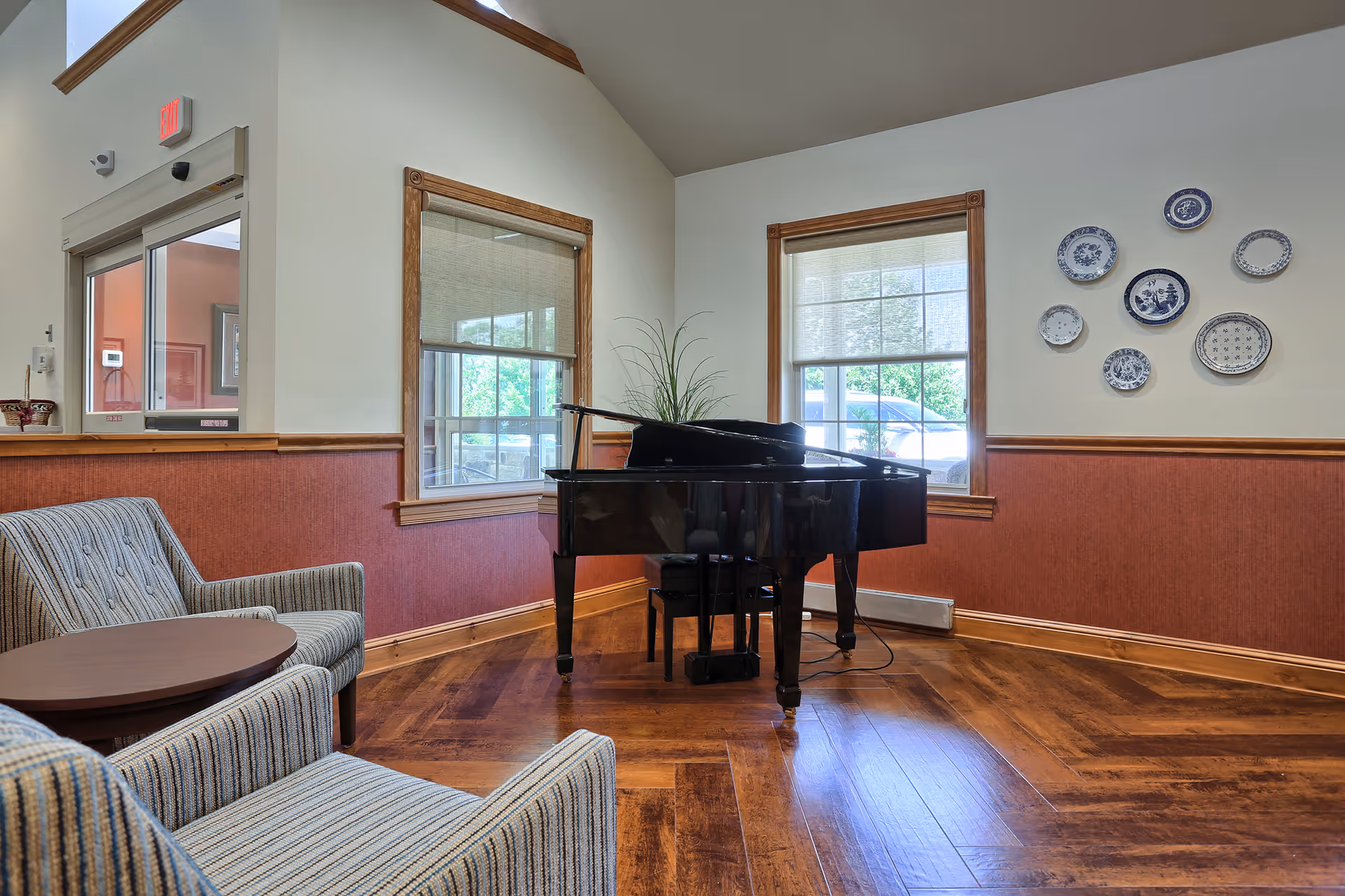 A cozy interior room with two striped armchairs and a small round wooden table in the foreground. A black grand piano with a microphone stand is positioned near two windows with wooden frames. Decorative blue and white plates are arranged on the wall to the right. The floor is wooden with a herringbone pattern, and the walls are painted white with a reddish-brown wainscoting.