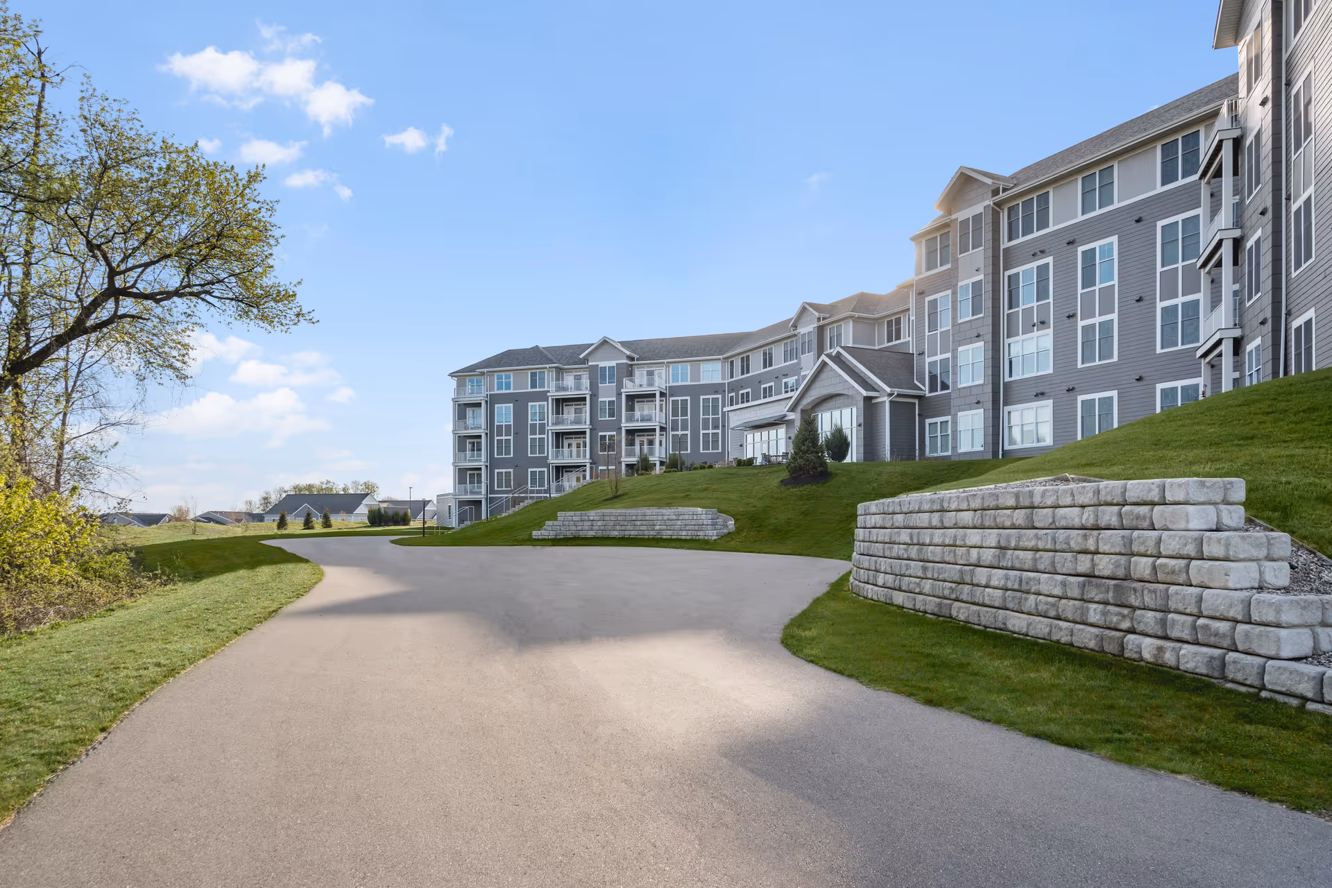 Exterior view of a multi-story senior living building with a curved road and stone walls.