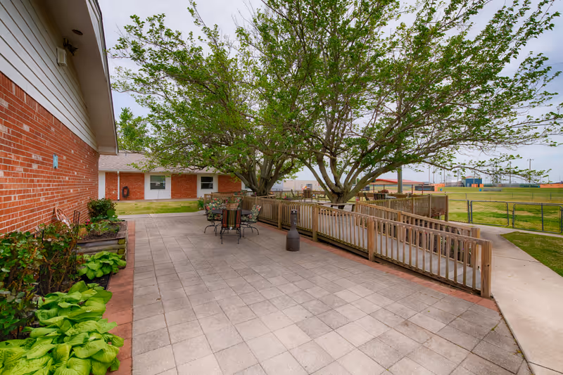 Outdoor patio area at Shawnee Colonial Estates featuring a paved surface with a table and chairs under large leafy trees. There is a wooden ramp with railings leading to the patio, a brick building on the left, and a grassy field with a fence and sports field in the background under a cloudy sky.