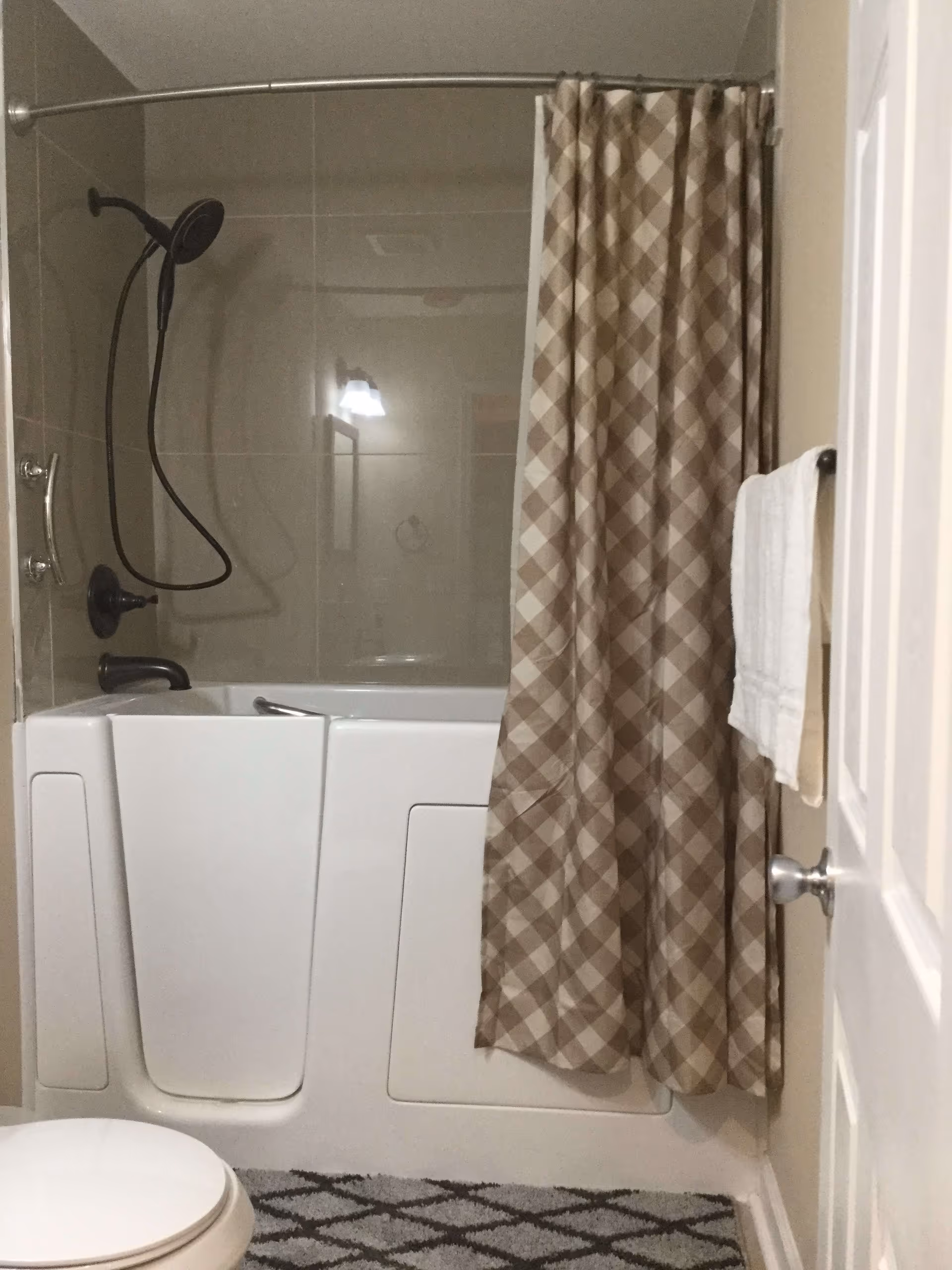A bathroom featuring a walk-in bathtub with a door, a handheld showerhead mounted on the wall, a beige and white checkered shower curtain, a white towel hanging on a towel rack, a toilet, and a patterned bath mat on the floor.