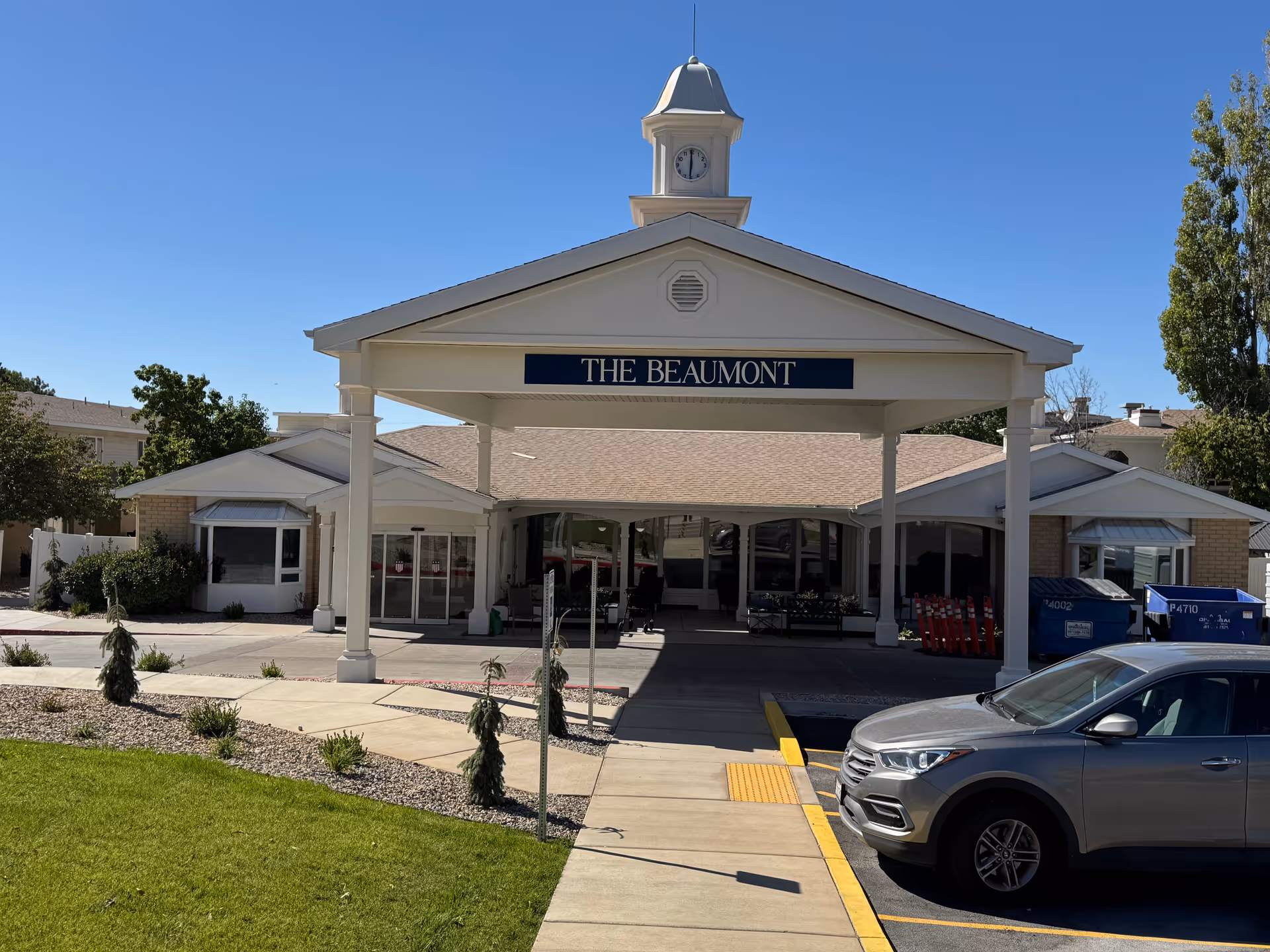 Front exterior view of The Beaumont Assisted Living and Memory Care building with a covered entrance, clock tower on top, and a parked silver SUV in front. The sky is clear and blue, with some landscaping and trees around the building.