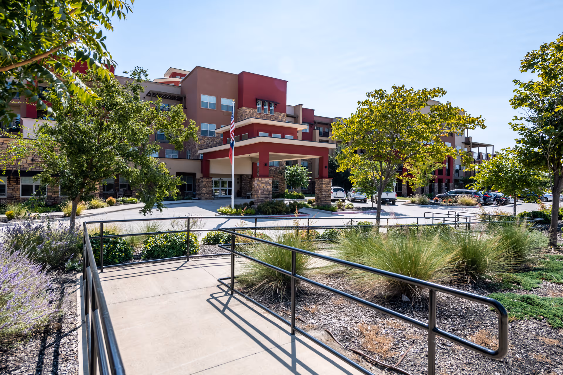 Exterior view of Alexis Estates Gracious Retirement Living building with a covered entrance, surrounded by trees, shrubs, and a paved walkway with handrails leading to the entrance. Several cars are parked in the parking lot on a sunny day.