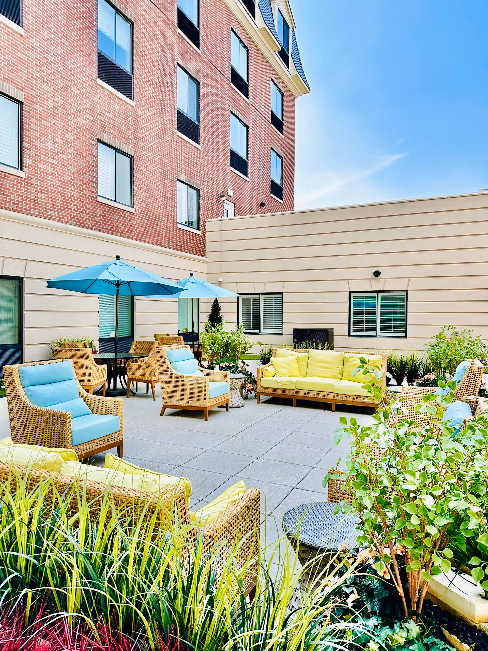Outdoor patio with wicker seating, blue cushions, umbrellas, and lush plants beside a brick building.