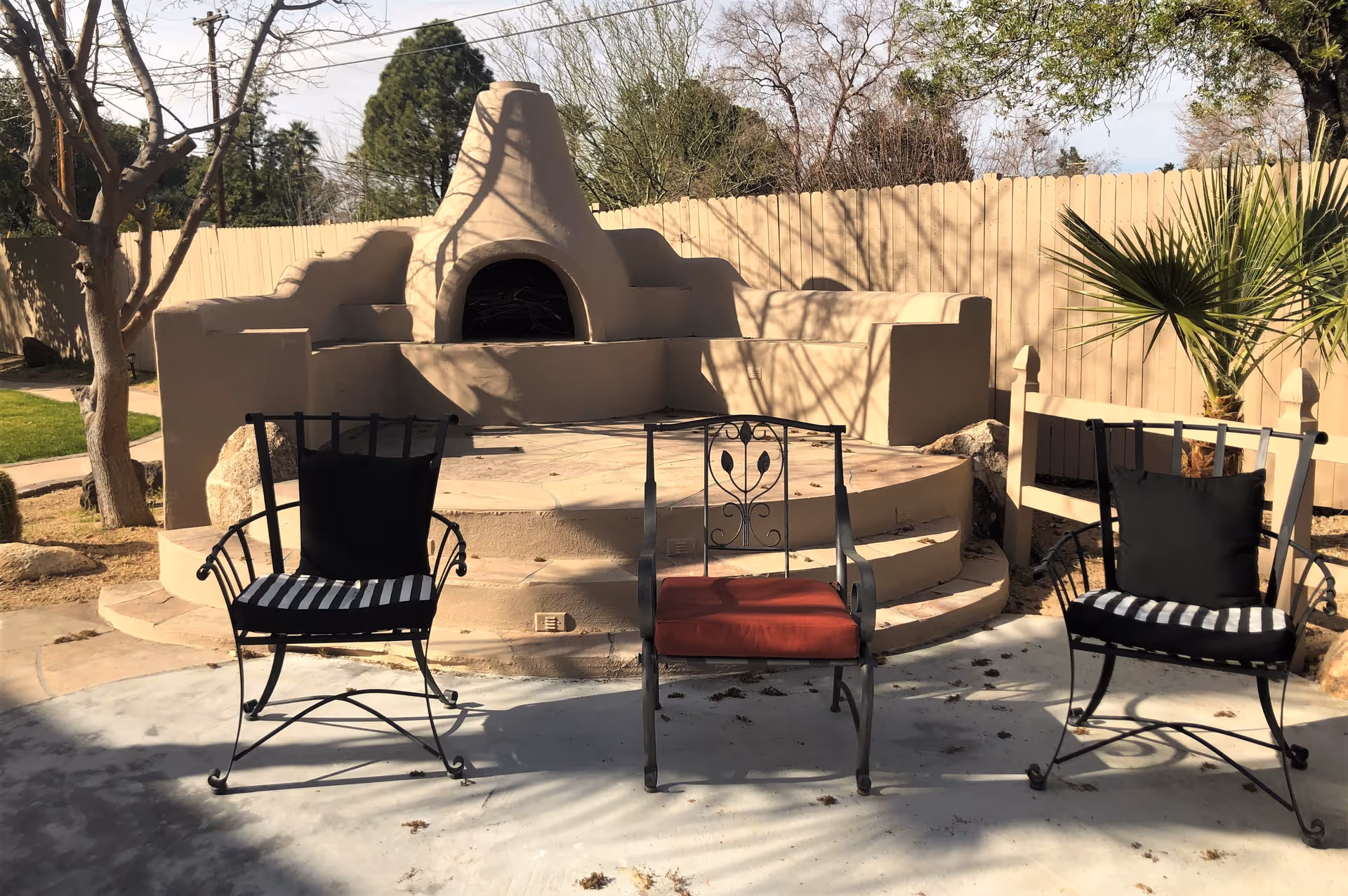 Outdoor patio area with three metal chairs, two with black and white striped cushions and one with a red cushion, arranged in front of a built-in adobe-style fireplace and seating area. The space is enclosed by a wooden fence with trees and plants visible in the background.