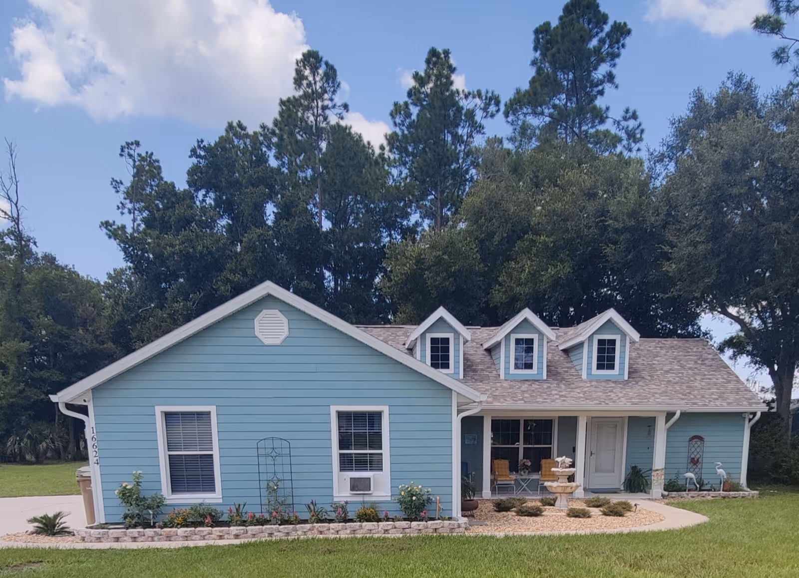 Blue single-story assisted living facility with a front porch, landscaping, and tall trees in the yard.