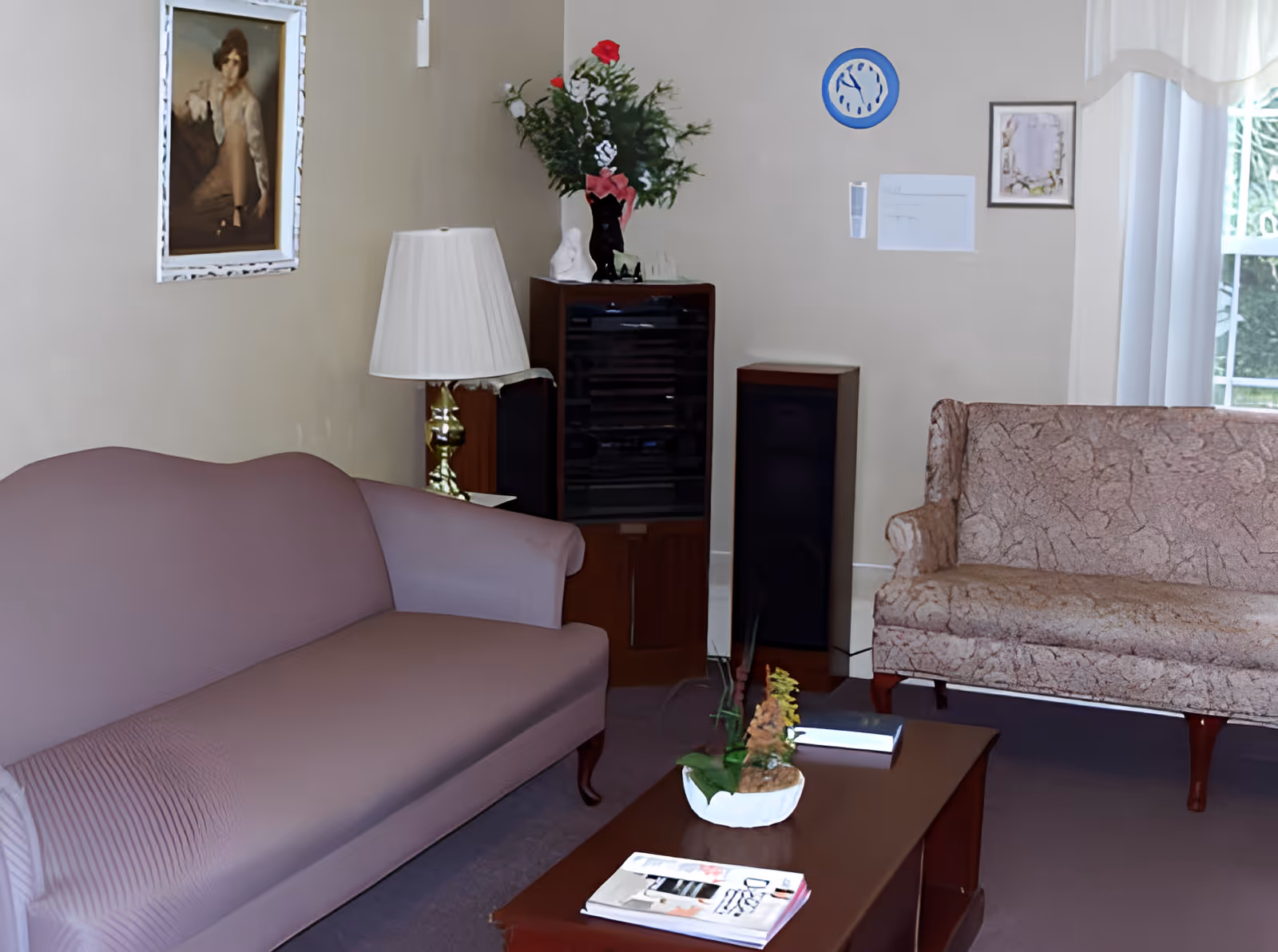 A cozy living room with two sofas, one purple and one beige patterned, a wooden coffee table with a plant and magazines, a floor lamp, a cabinet with a vase of flowers, a speaker, a blue clock on the wall, and framed pictures. A window with sheer white curtains lets in natural light.