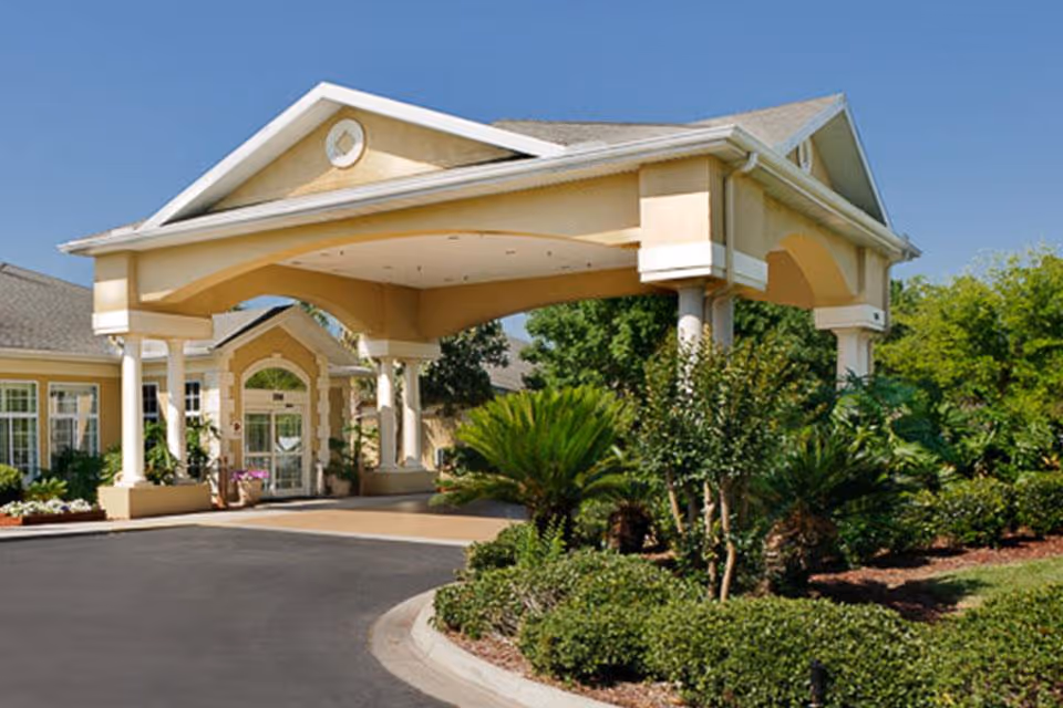 Front exterior view of a senior living facility with a covered entrance supported by white columns, surrounded by well-maintained landscaping including bushes and small trees under a clear blue sky.