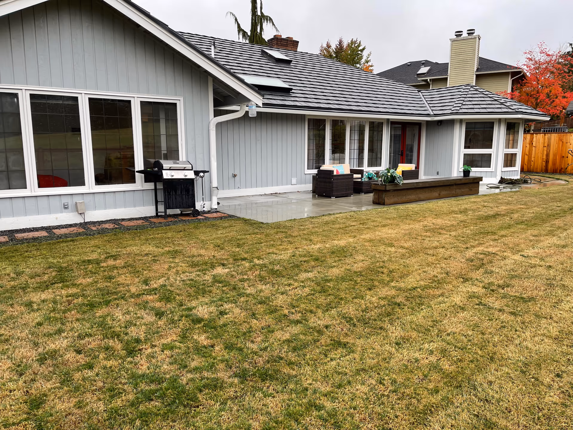 Backyard view of a single-story gray house with a patio area featuring outdoor seating and a barbecue grill. The yard has a large grassy lawn and a wooden planter box with plants. Trees with autumn foliage are visible in the background.