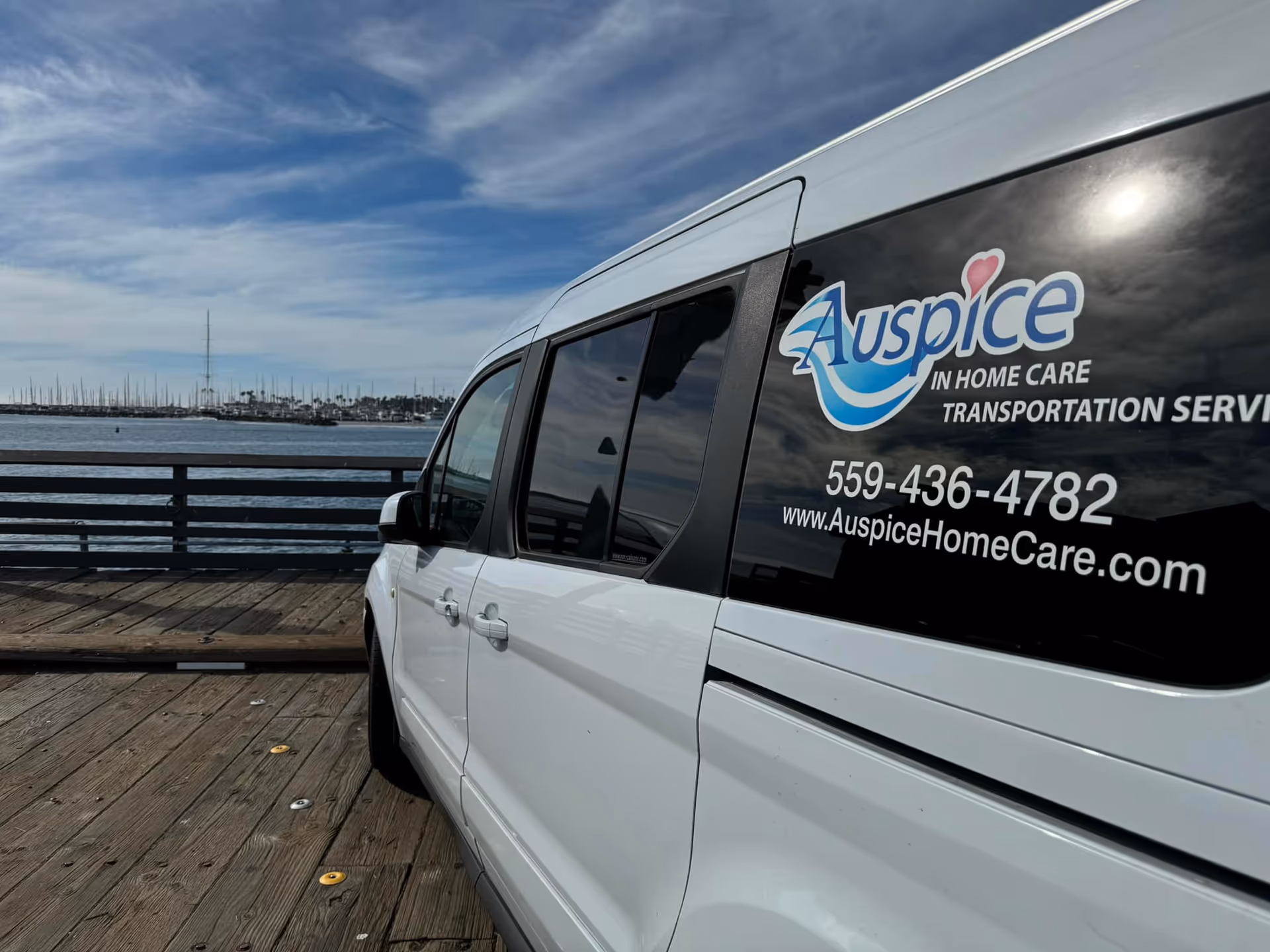 White Auspice transportation van with logo and phone number parked on a wooden boardwalk.