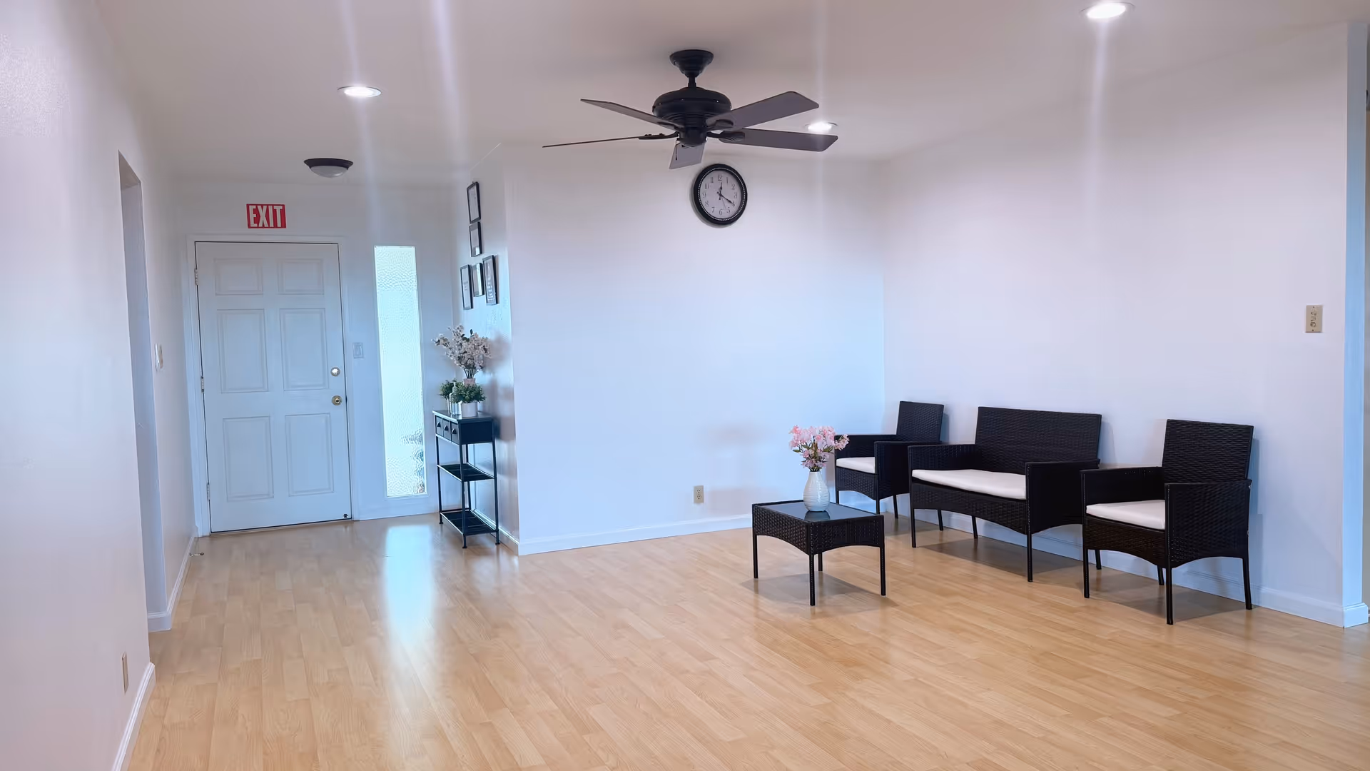 Bright lobby with wicker seating, a coffee table, clock, ceiling fan, and exit door.