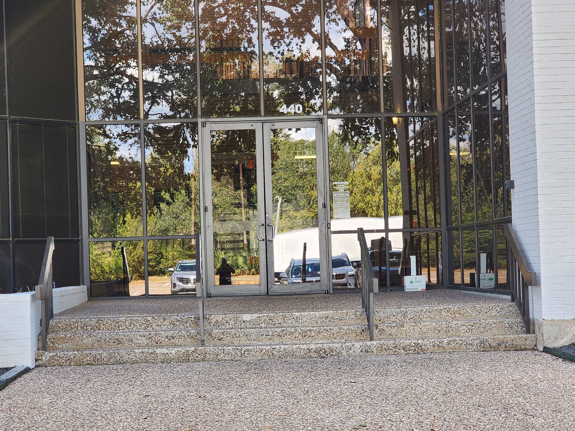 Glass-front entrance with metal doors, steps, and reflections of trees.