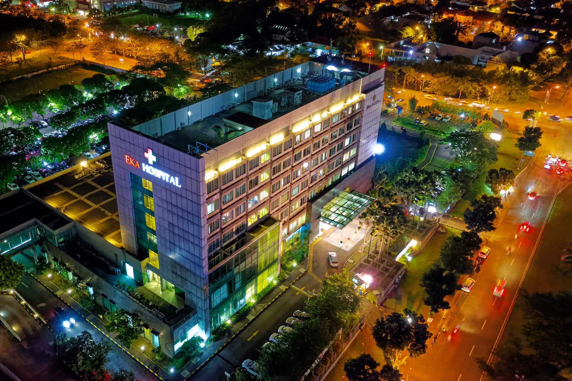 Aerial night view of a multi-story hospital with illuminated signage beside a tree-lined road.