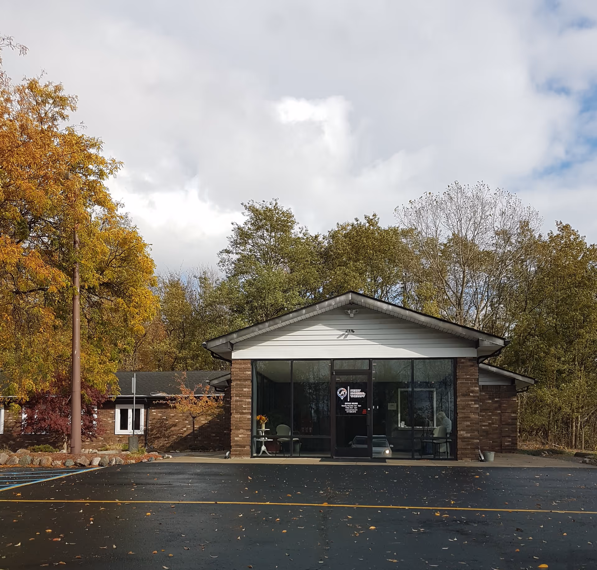 Small brick clinic building with glass entrance, surrounded by autumn trees and a paved parking lot.