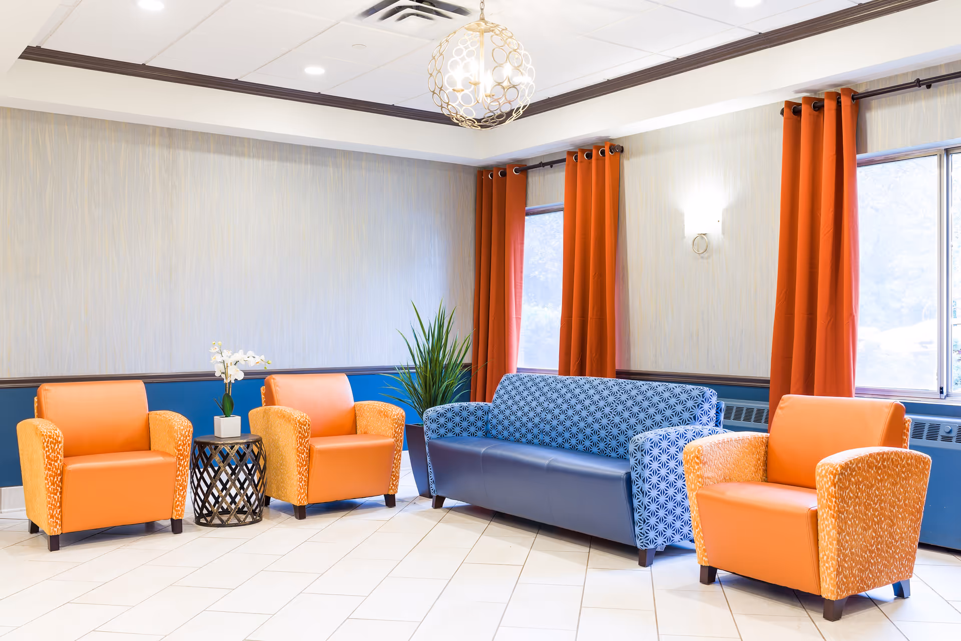A bright and clean seating area with three orange armchairs and one blue patterned sofa arranged around a small black side table with a white orchid plant. The room features light-colored walls with a blue lower trim, orange curtains on the windows, and a modern spherical ceiling light fixture.