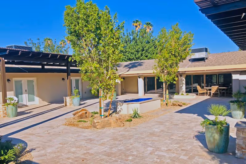 Sunny courtyard with a central fountain, potted plants and young trees surrounded by a one-story building and outdoor seating.