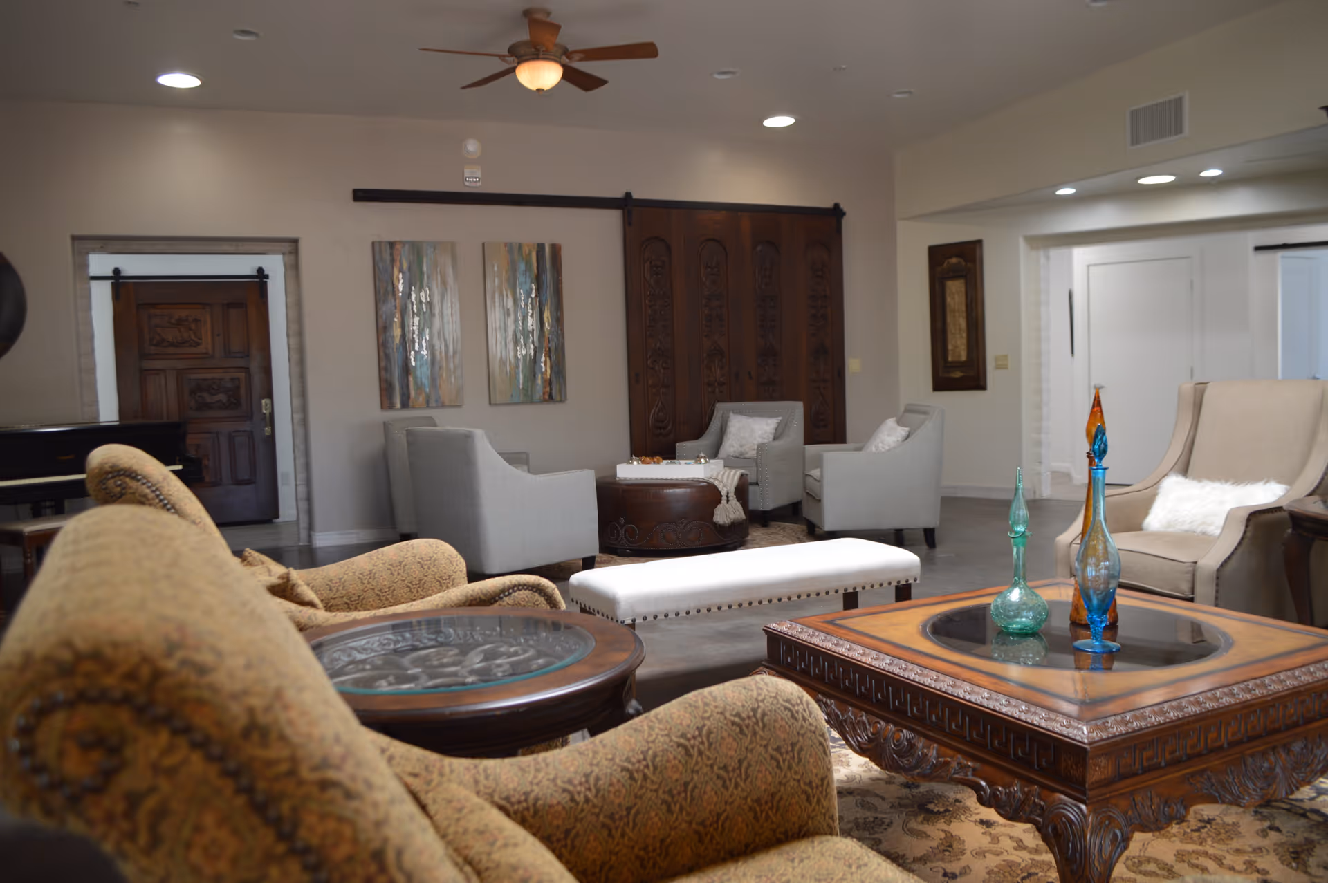 A spacious and well-furnished living room in an assisted living facility featuring multiple upholstered chairs and sofas arranged around wooden coffee tables with decorative glass vases. The room has neutral-colored walls, a ceiling fan with light, and decorative wall art. There is a wooden door and a piano visible in the background.