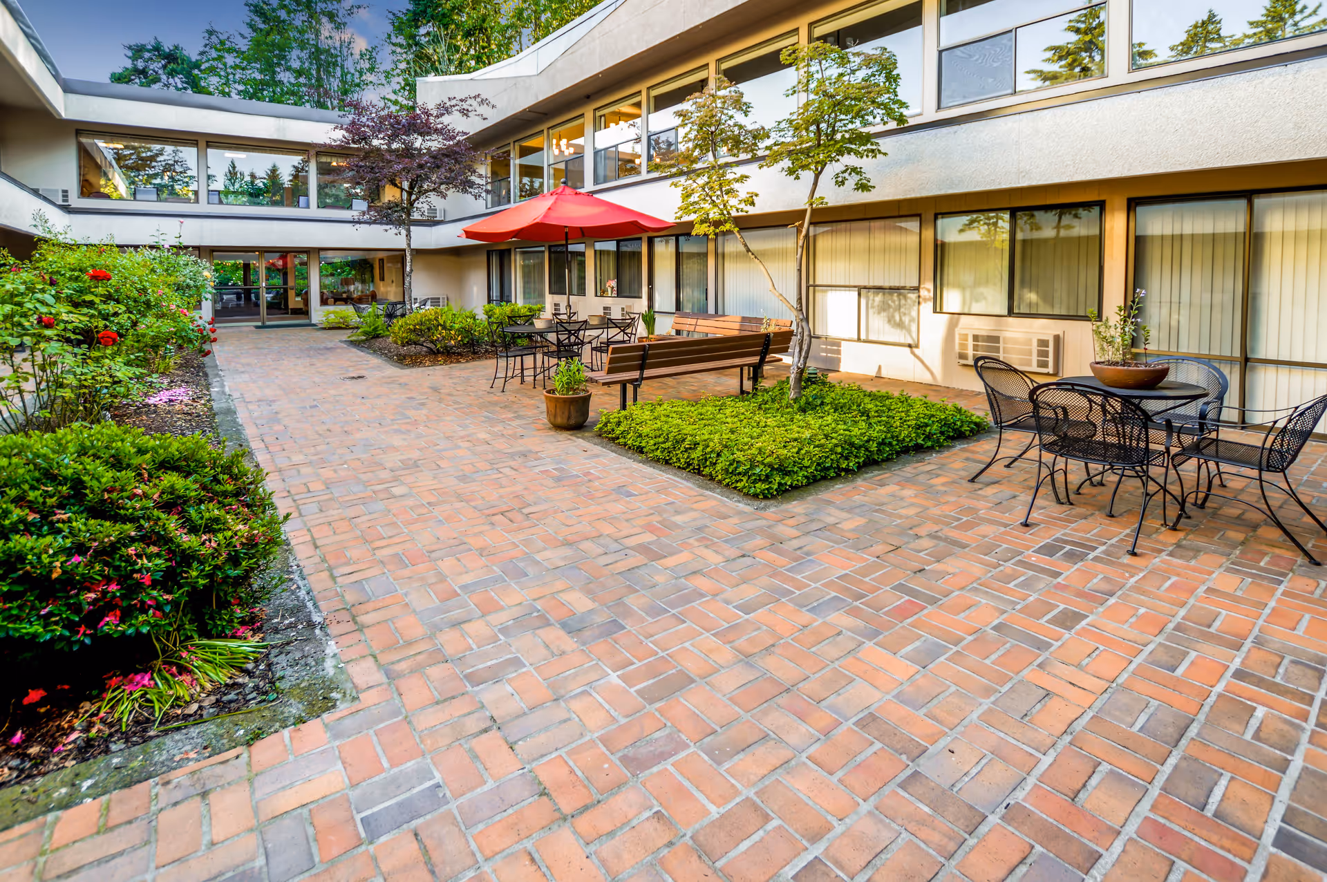 Outdoor courtyard area of an assisted living facility with brick flooring, garden beds with shrubs and flowers, metal tables and chairs, wooden benches, and a red umbrella providing shade. The building has large windows and is surrounded by trees.