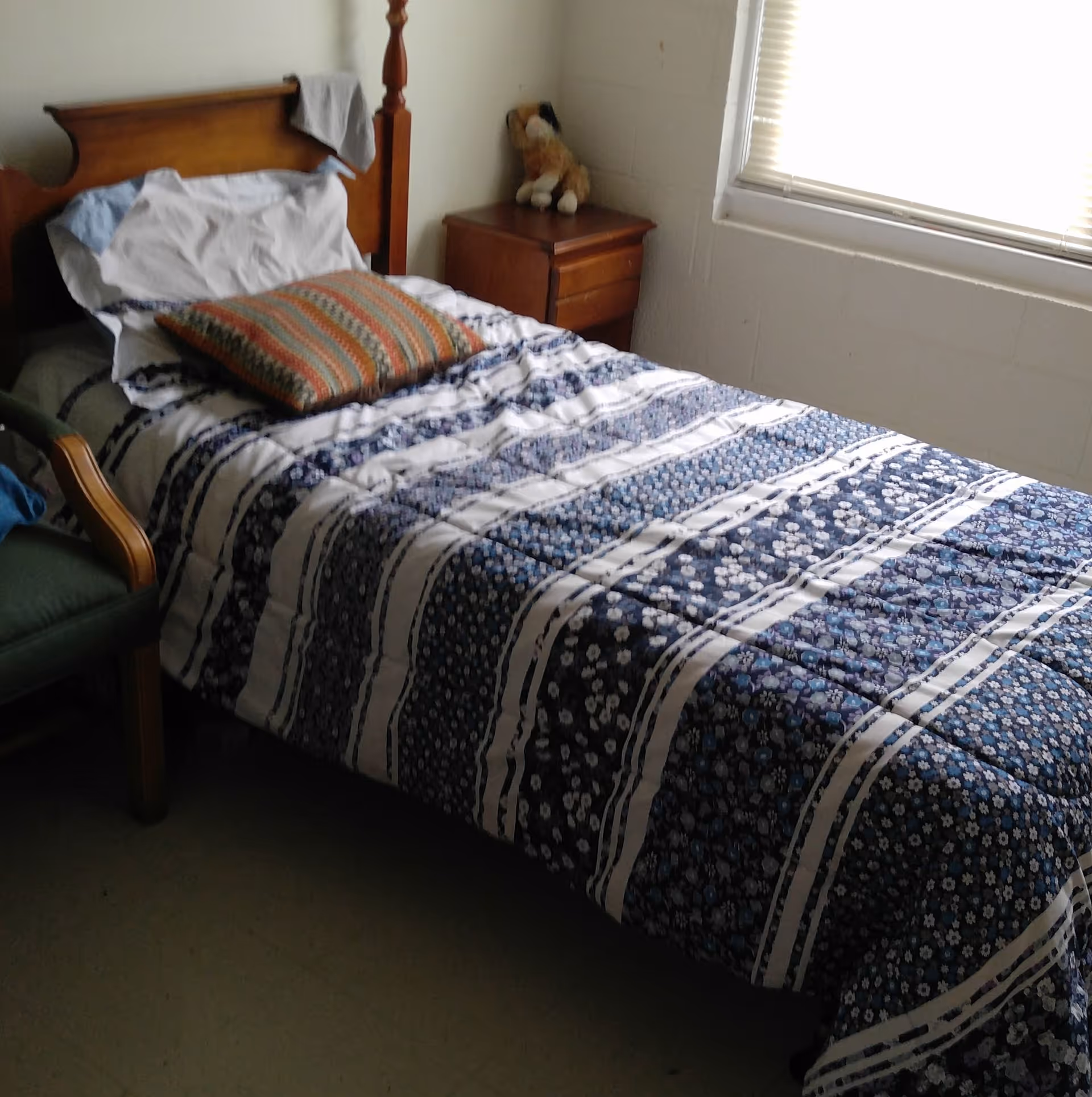 A small bedroom with a single bed covered in a blue and white floral patterned comforter, a striped pillow, and a white pillow. Next to the bed is a wooden nightstand with a stuffed dog toy on top. A window with closed blinds is on the right wall, and a green armchair is partially visible on the left side.
