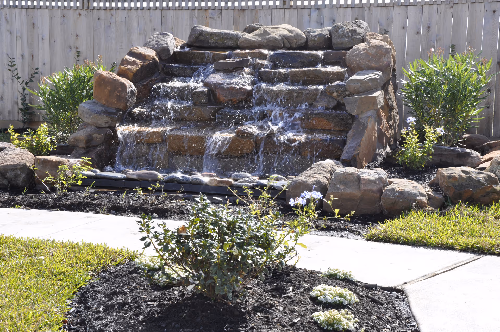 Outdoor rock waterfall fountain with shrubs and a wooden fence in a landscaped yard.