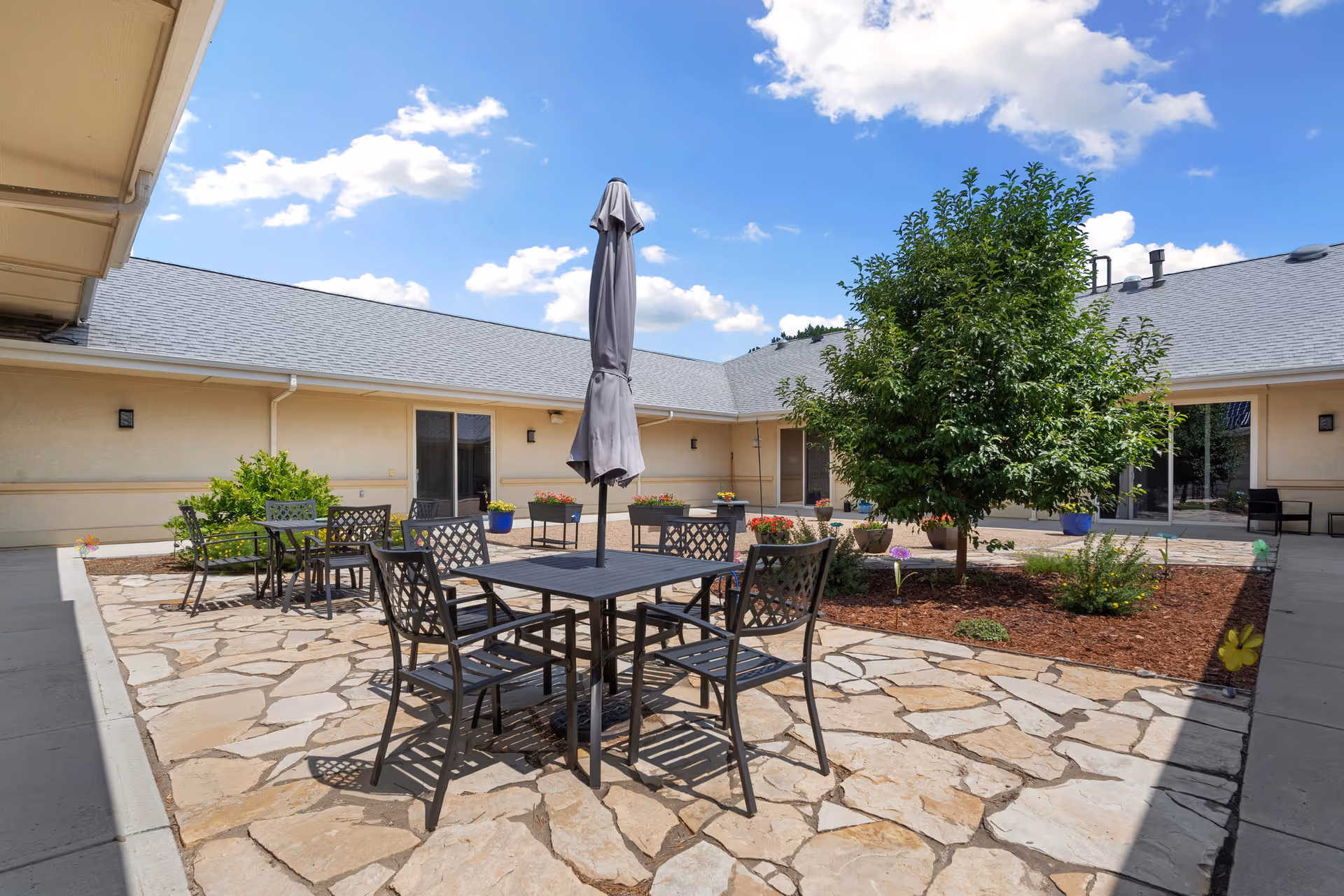 An outdoor courtyard with stone patio, tables, chairs, a central umbrella, and greenery.