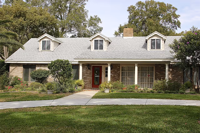 Brick senior living facility with a front porch, dormer windows, and a well-kept lawn.