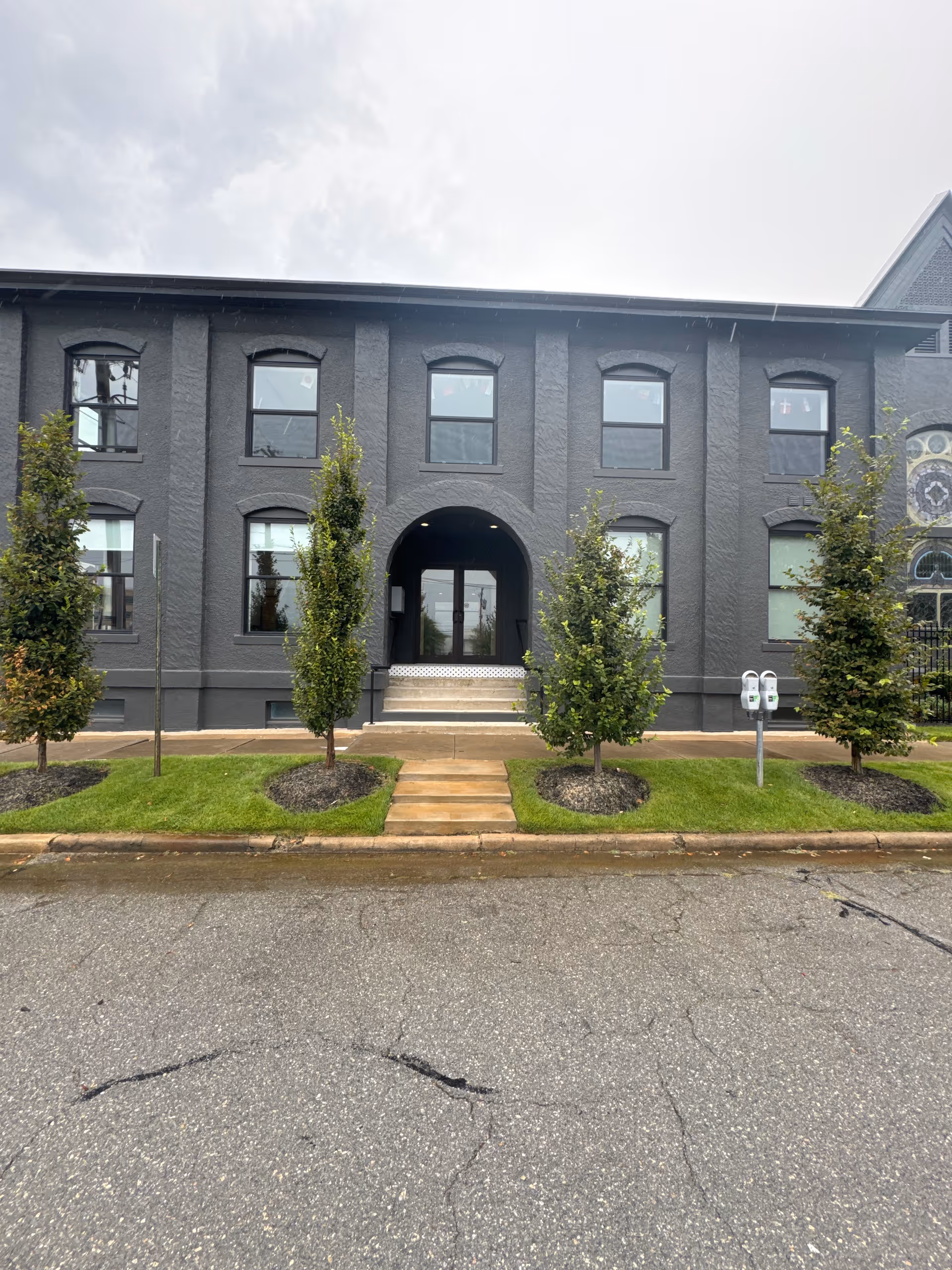 Dark gray two-story building with an arched entrance, steps, and small trees along a sidewalk.