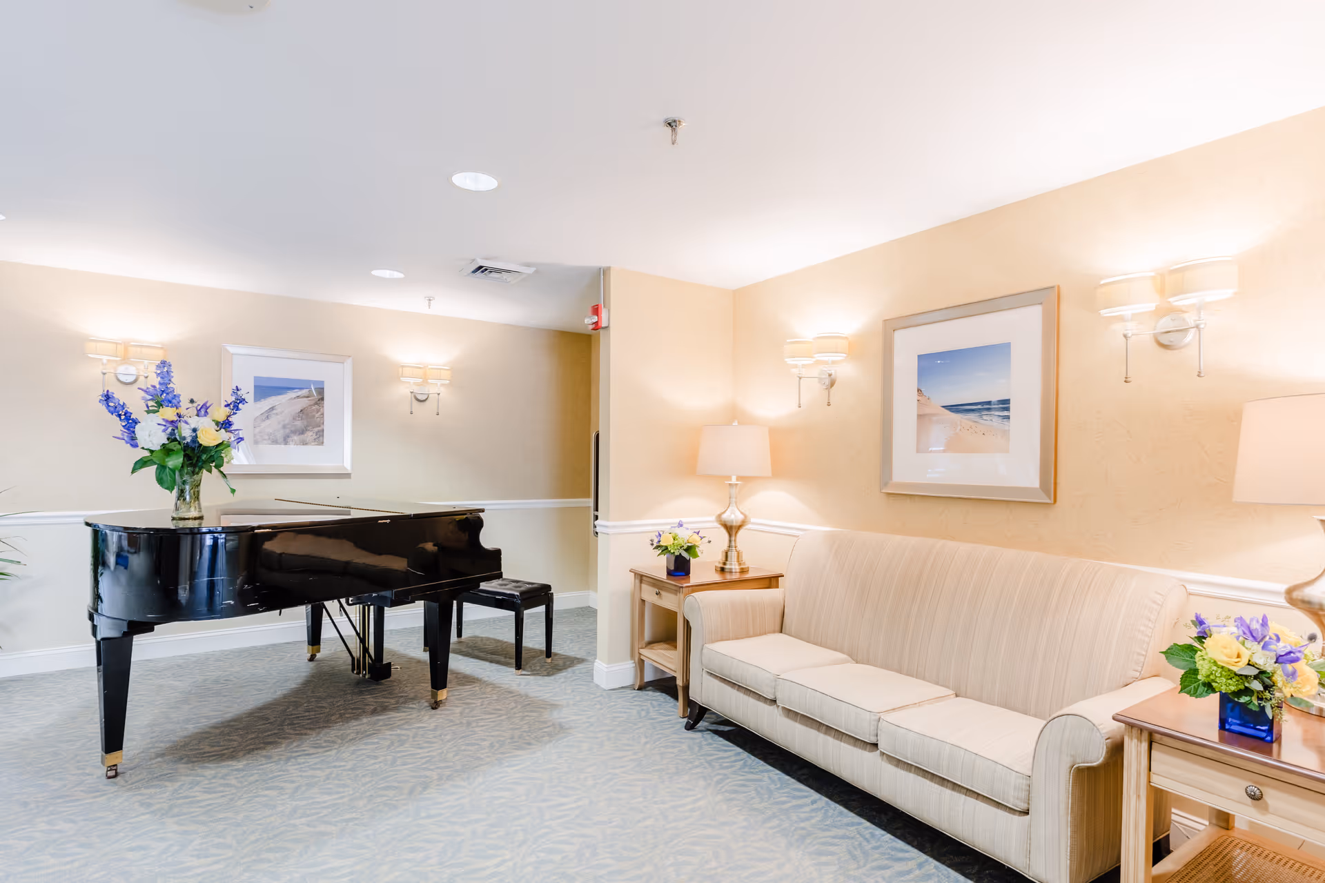 Bright sitting room with a black grand piano, a beige sofa, side tables with lamps and floral arrangements, and framed seaside artwork.