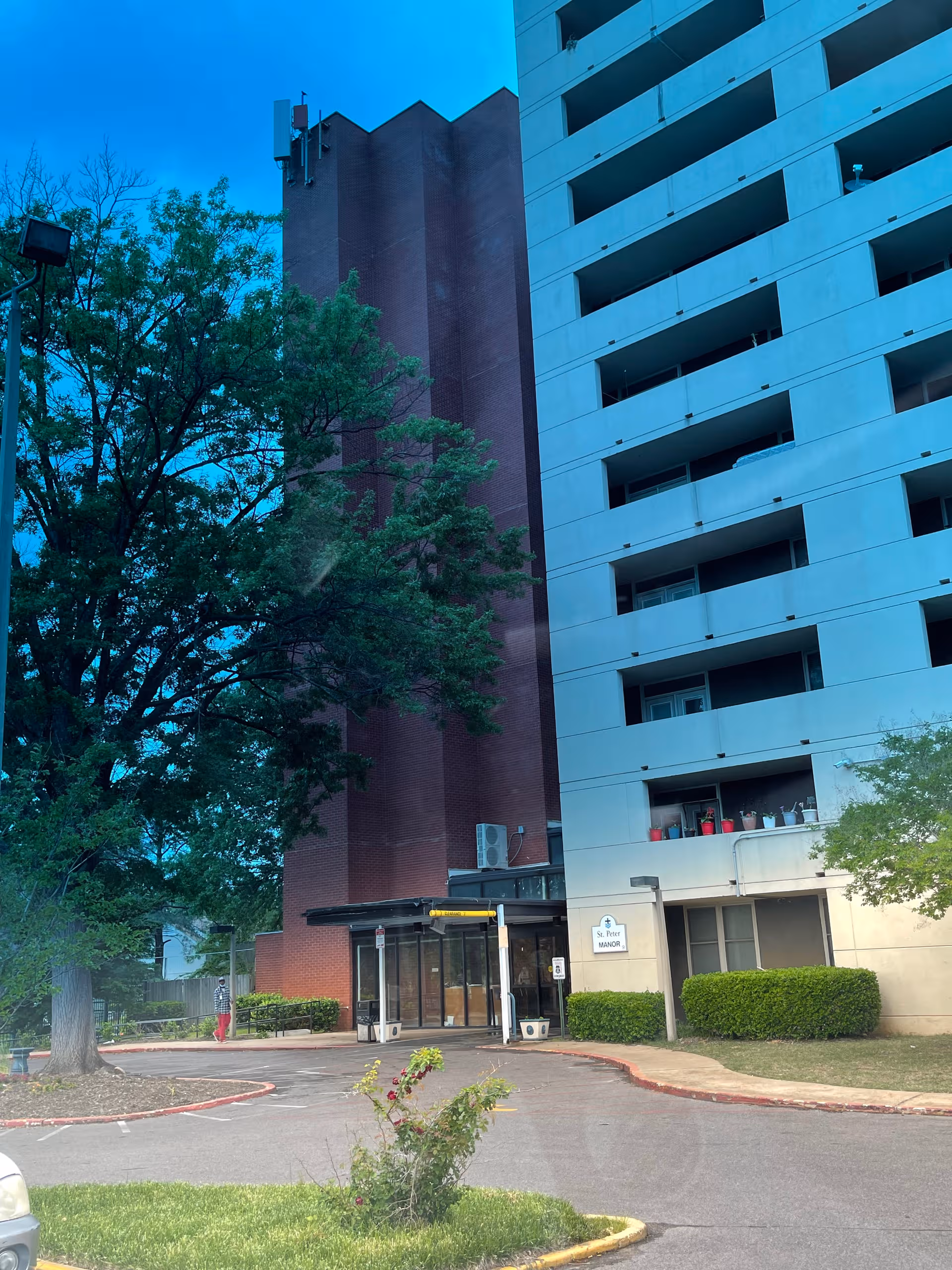 Tall multi-story senior living building labeled St. Peter Manor with glass entrance and trees.
